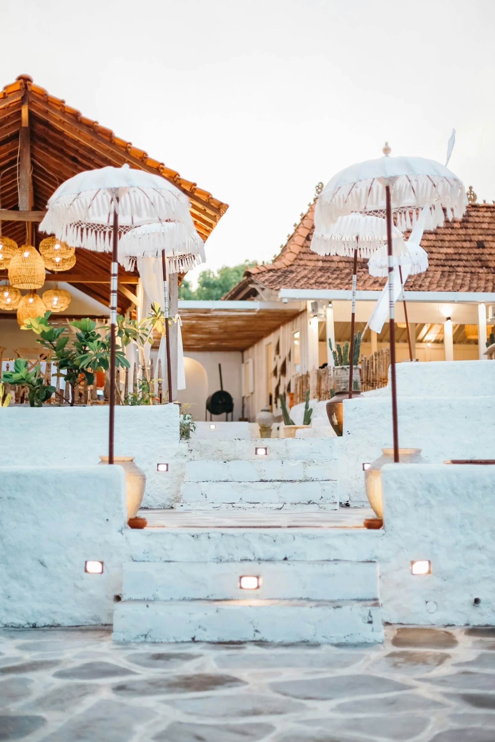 White steps with built-in lighting lead up to an outdoor patio area with white umbrellas and potted plants, featuring a building with traditional tiled roof and hanging lanterns.