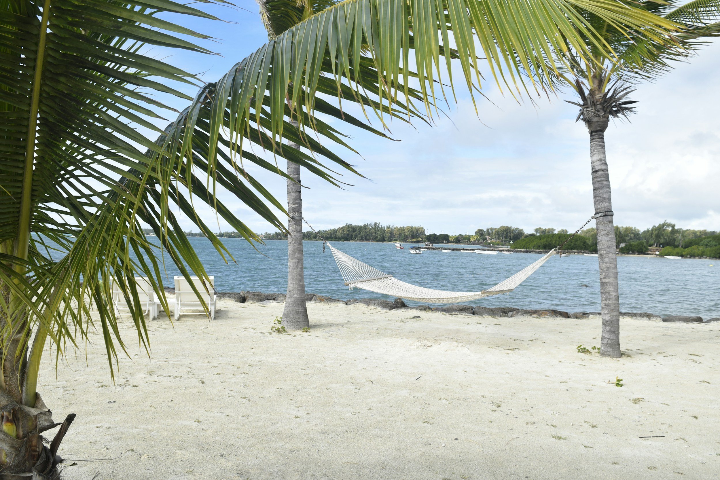 A tropical beach scene with a hammock hanging between two palm trees, white sand, lounge chairs, and a view of water with boats and a distant shoreline under a partly cloudy sky.