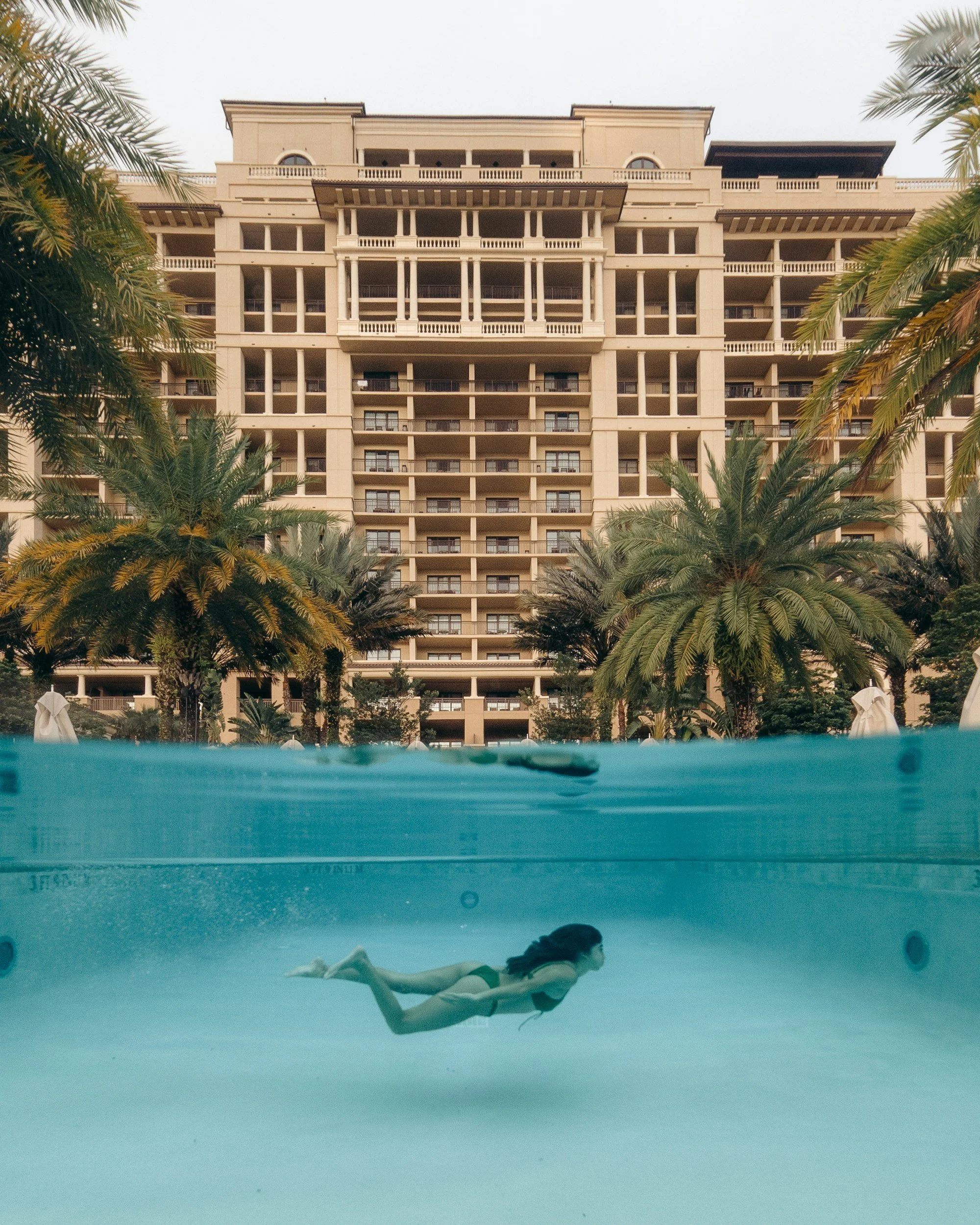 A woman swimming underwater in a pool with a large beige building and palm trees in the background.
