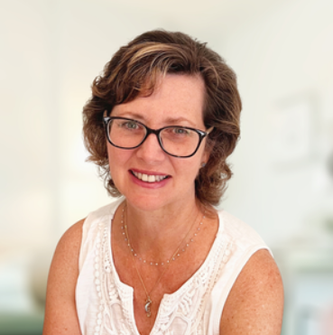 A woman with short wavy brown hair and glasses, smiling, wearing a white sleeveless top.