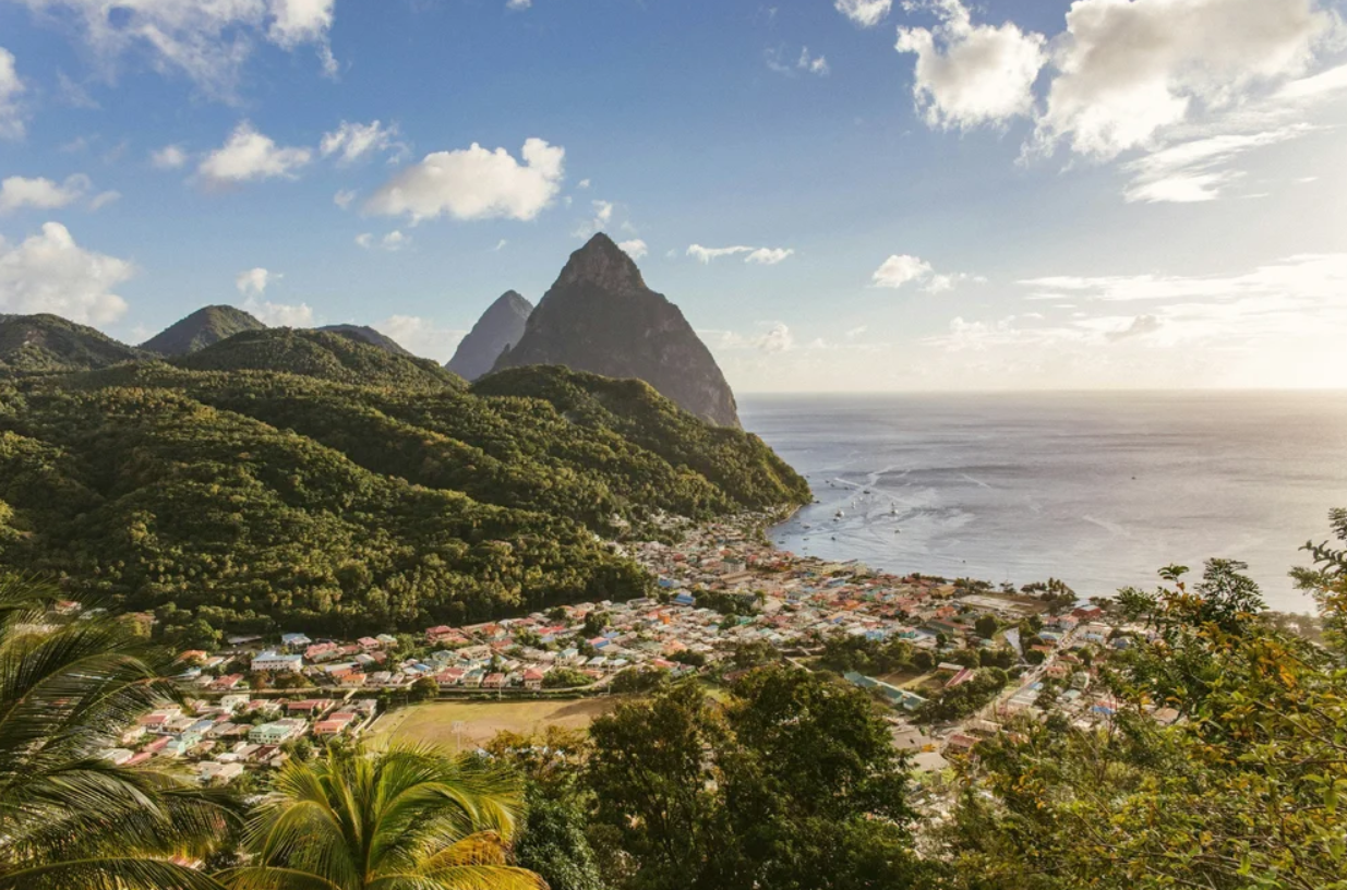 Scenic coastal landscape featuring lush green hills, a small town with colorful buildings, and a mountain with the ocean in the background under a partly cloudy sky.