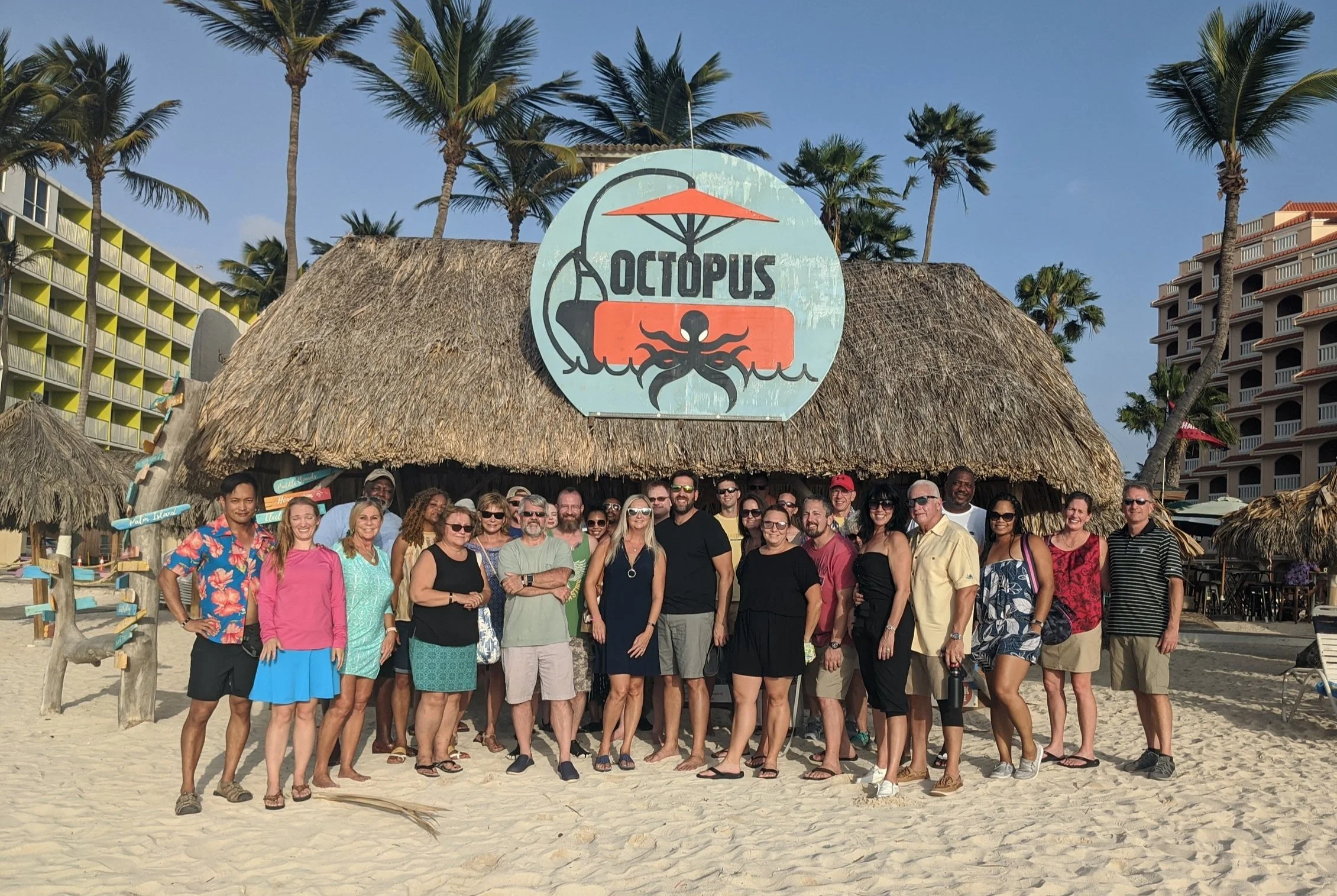 Group of people standing on a sandy beach in front of a thatched roof tiki bar with a sign that reads 'Octopus' with a picture of an octopus and a beach umbrella, palm trees, and hotel buildings in the background on a sunny day.