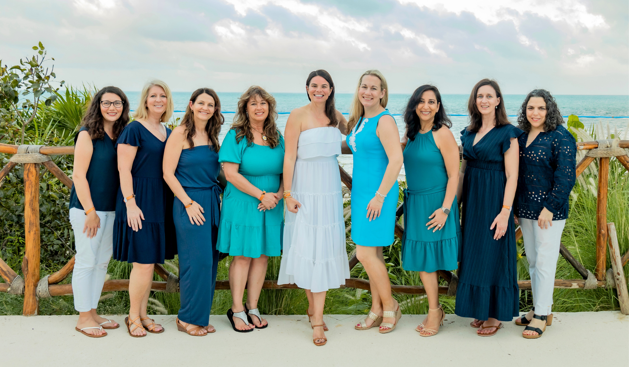 Group of nine women standing outdoors by the ocean, dressed in blue and white summer dresses, smiling for a photo.