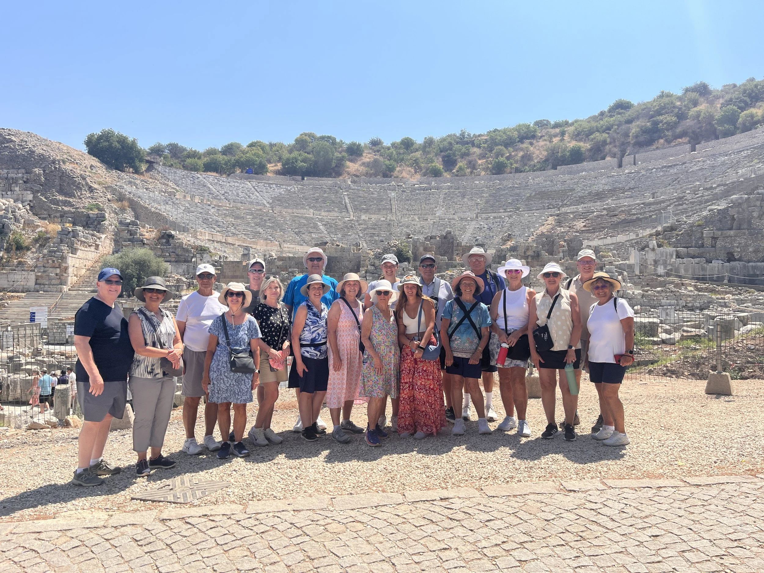 Group of tourists standing in front of ancient Greek ruins, possibly a historical amphitheater, on a sunny day with clear blue sky.