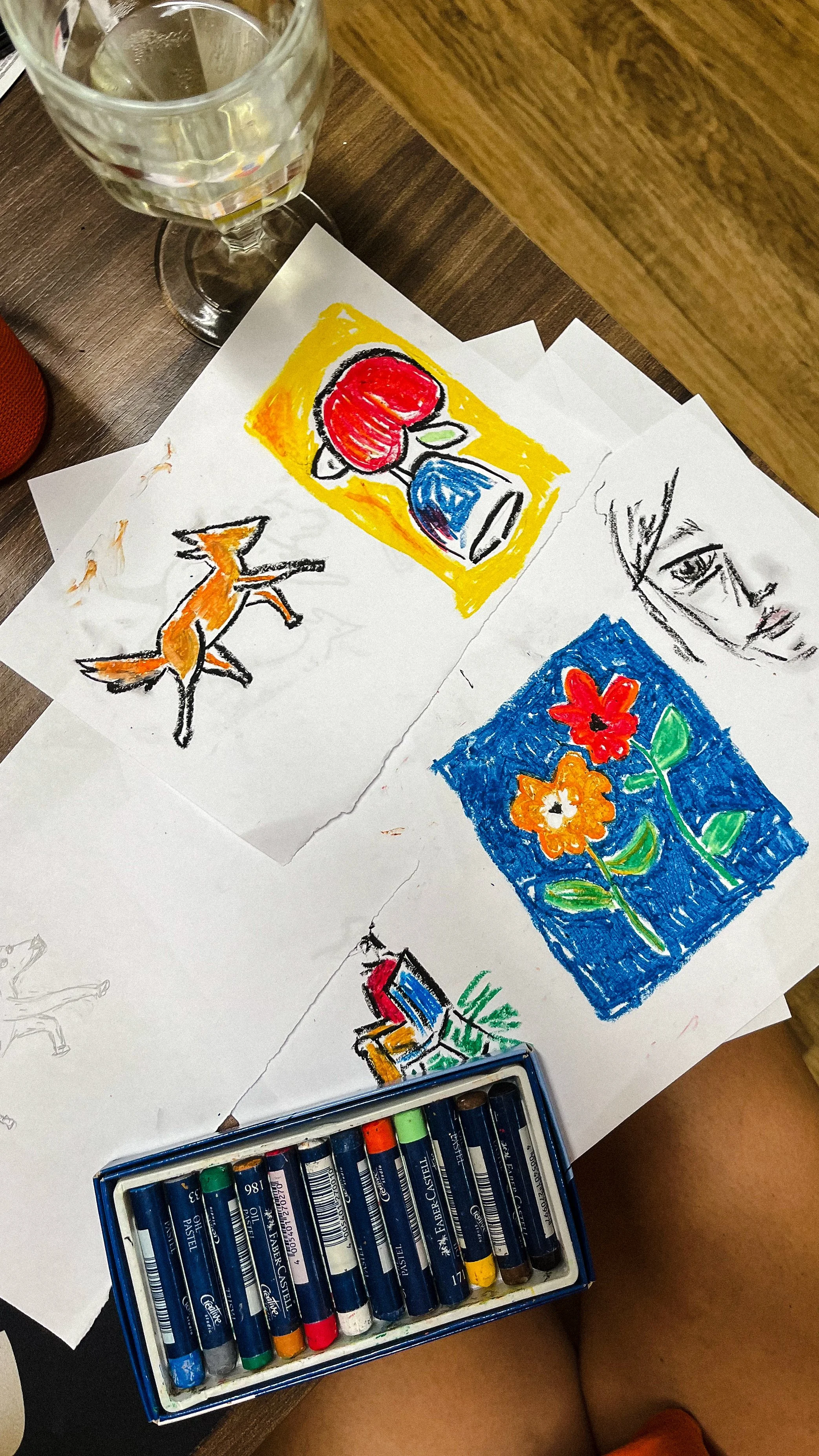 Colorful sketches on paper including a red apple with a blue vase, a fox, a woman's face, and two flowers, with a box of colored pastels in the foreground and a glass of water nearby.