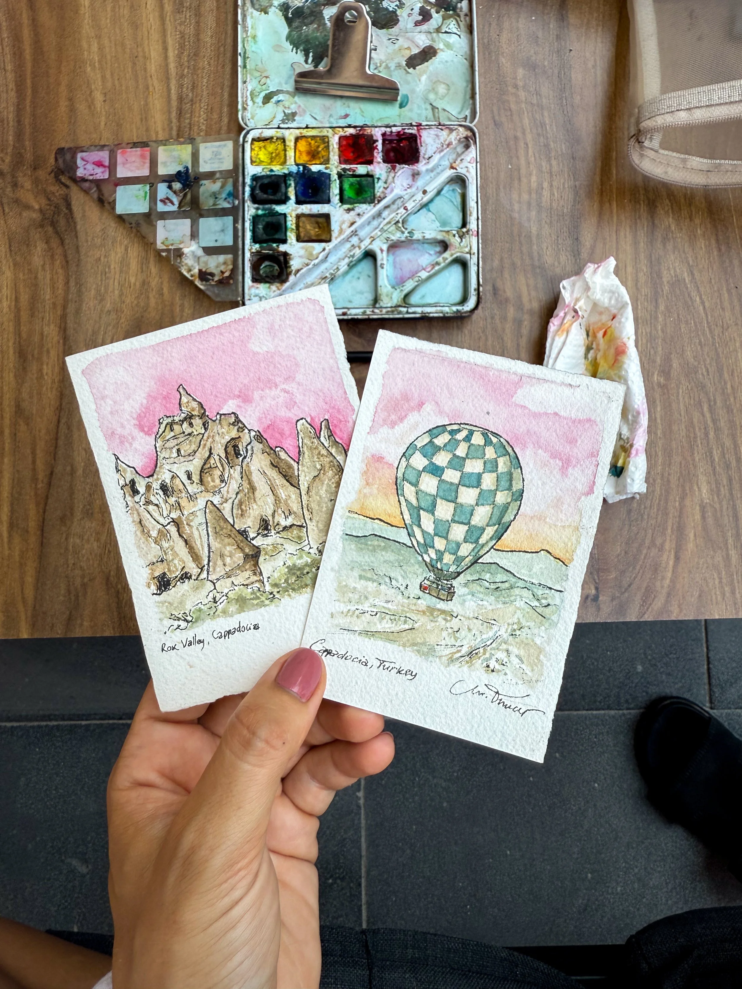 Two watercolor postcards featuring landscapes, one of Rox Valley Cappadocia, Turkey, with pink sky and rocky formations; the other of a hot air balloon over a landscape with pink sky.
