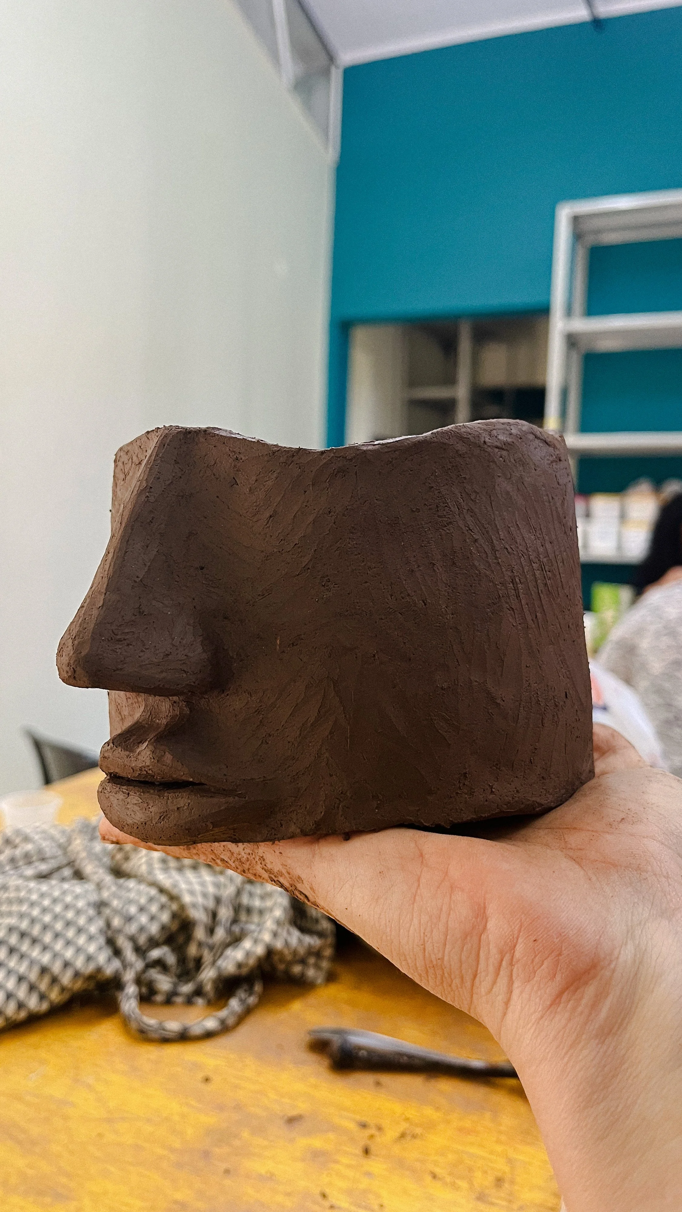 A hand holding a carved block of clay shaped like a human face in profile with prominent nose and lips, against an indoor workshop background.