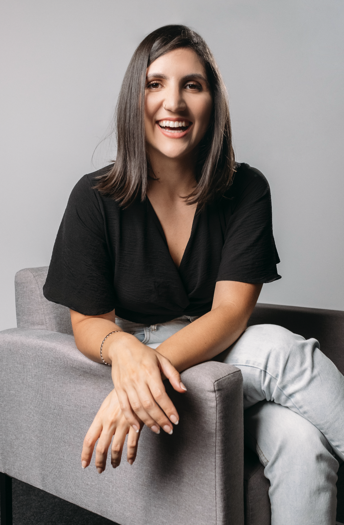 A woman with shoulder-length dark hair smiling, wearing a black top and light-colored pants, sitting on a gray armchair against a plain light gray background.
