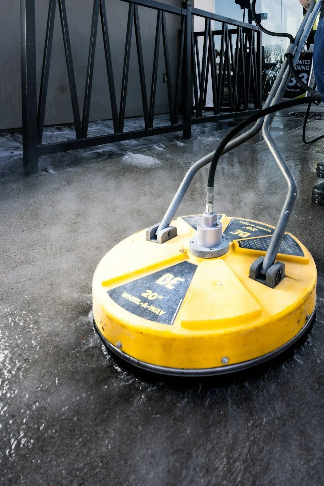 A yellow sidewalk scrubbing machine cleaning a wet concrete sidewalk outside a building.
