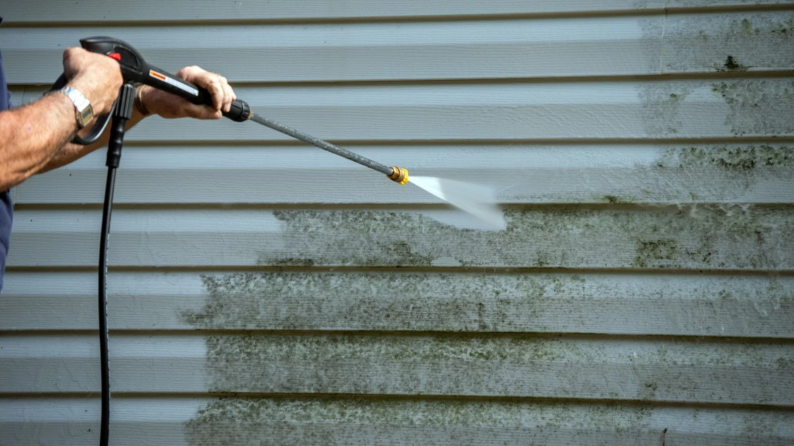 Person using a pressure washer to clean mold and dirt off a vinyl siding wall.