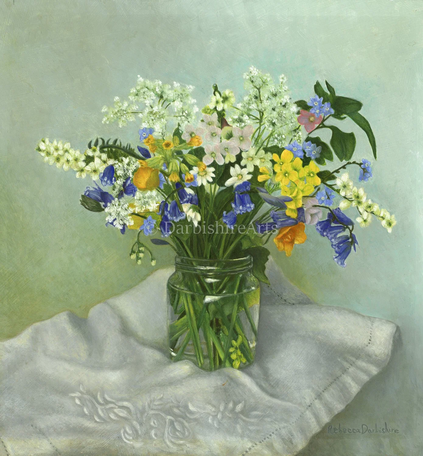 A glass jar filled with a colorful bouquet of wildflowers, placed on a white embroidered cloth against a light green background.