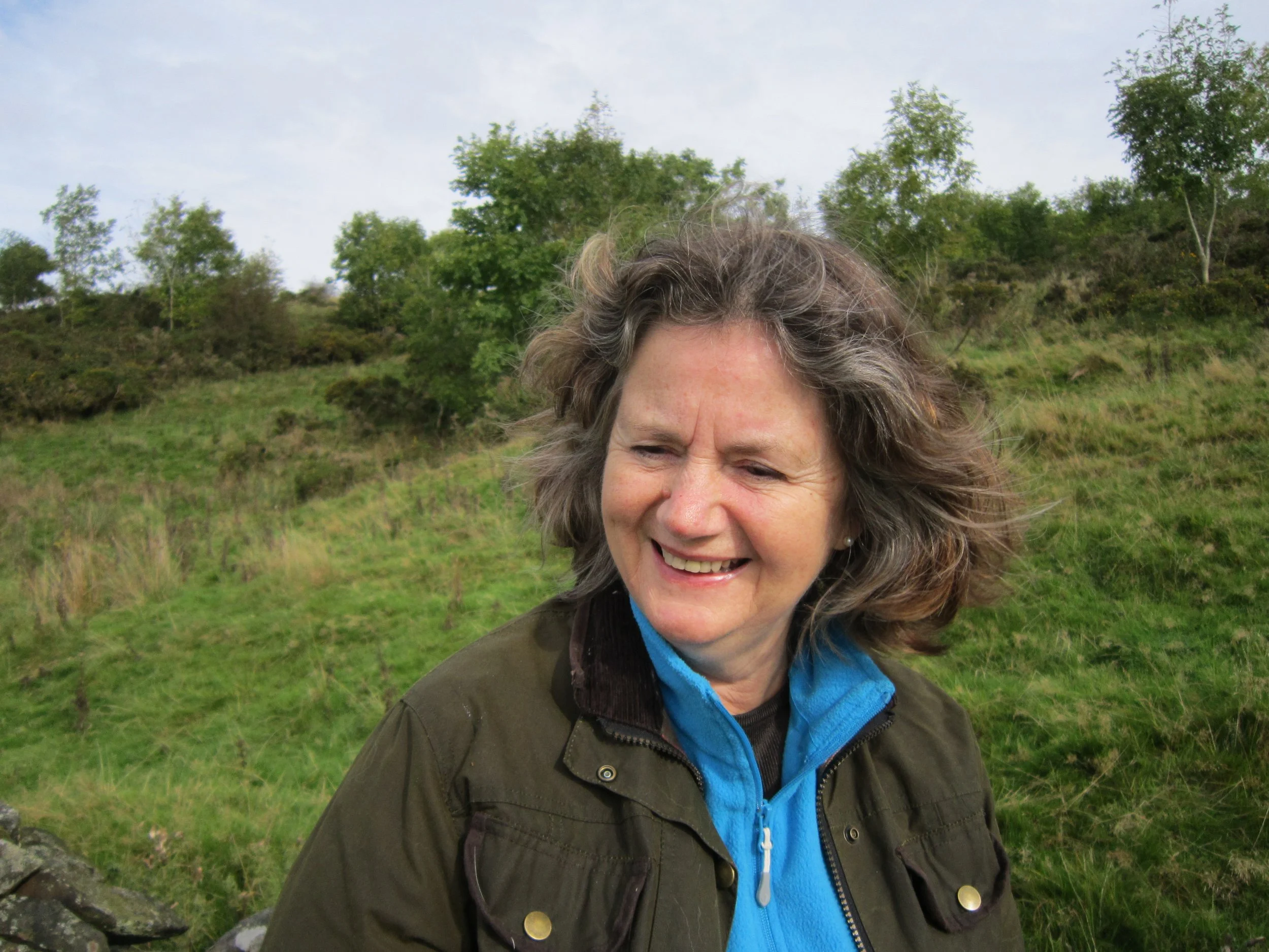 A woman with gray hair and a blue jacket smiling outdoors in a green, hilly landscape with trees.