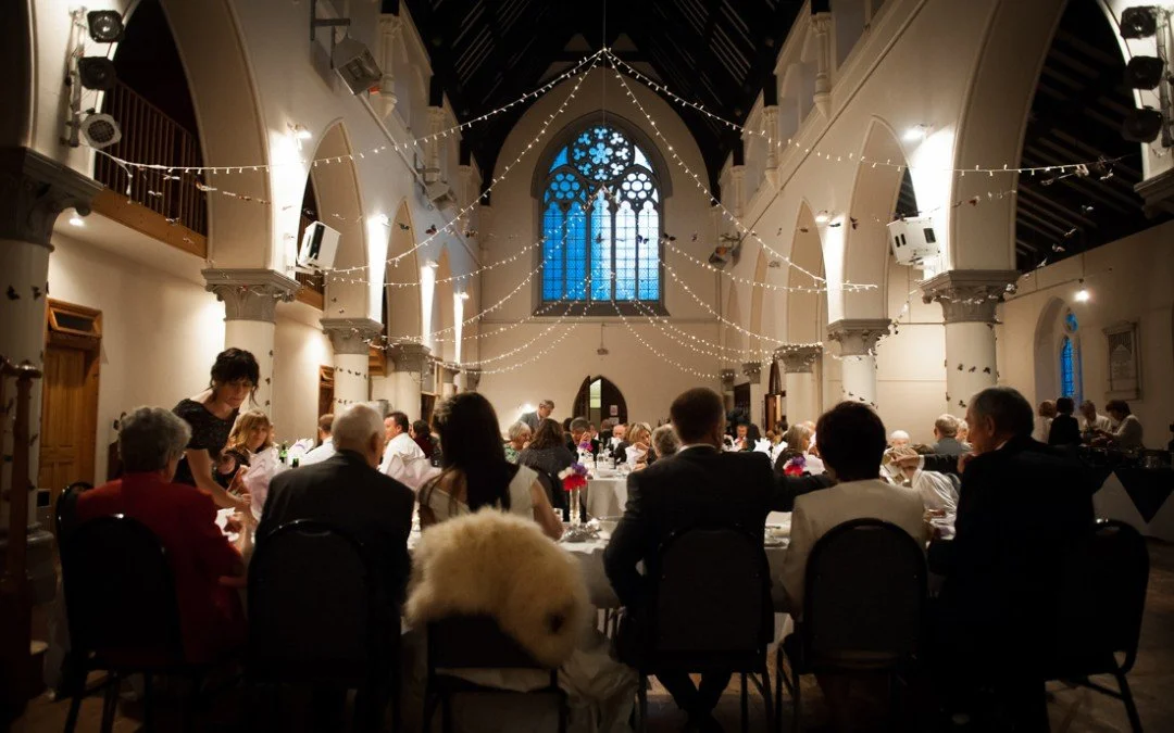 People seated at tables in a decorated banquet hall with high vaulted ceilings, stained glass windows, and string lights.