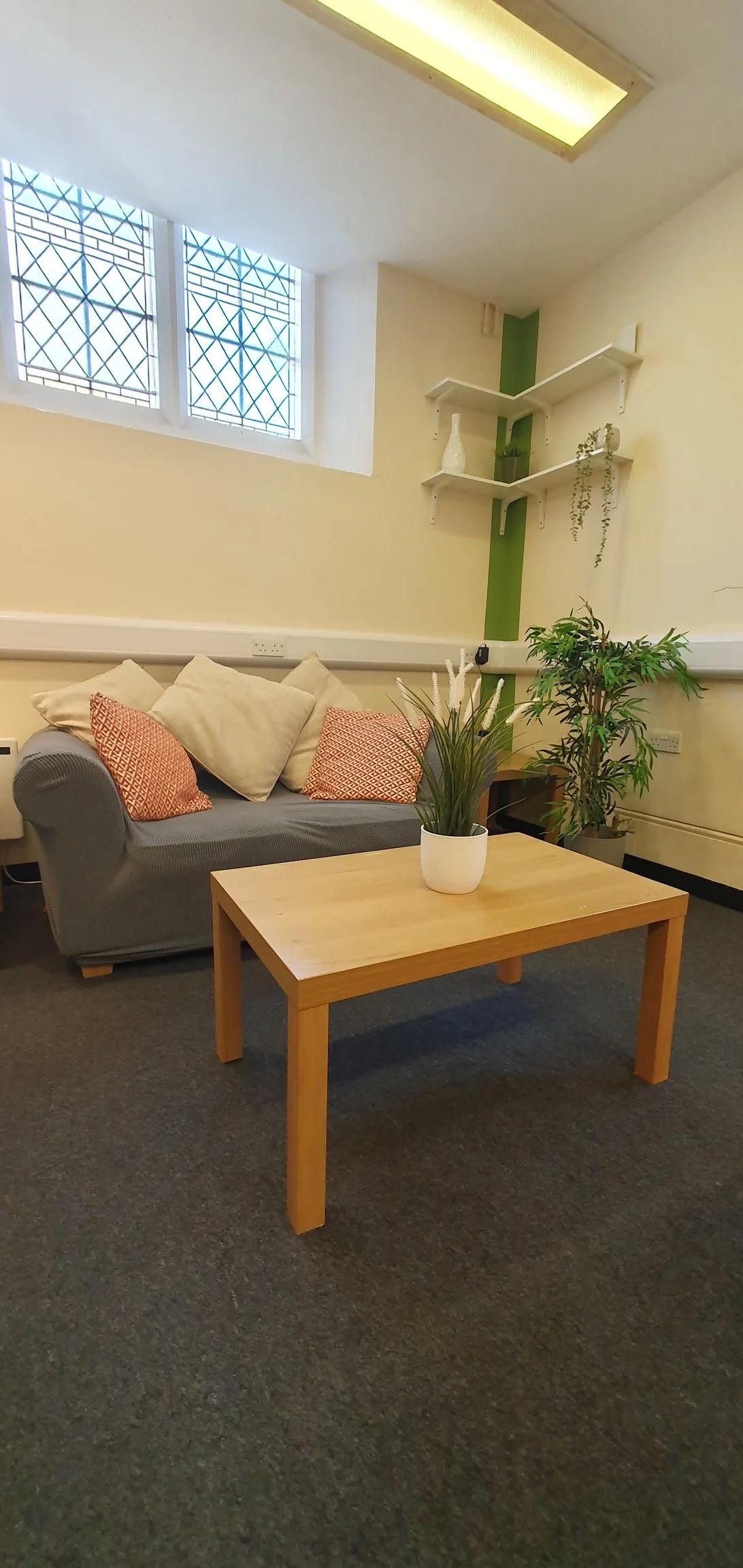 A cozy office or lounge area with a gray sofa decorated with beige and orange patterned pillows. A light wooden coffee table with a potted plant on top. Behind the sofa, large windows with diamond-patterned metal grilles let in natural light. Two whi