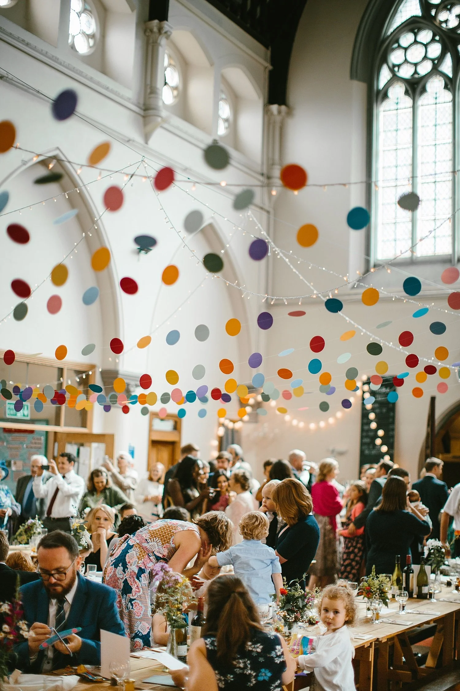 A lively indoor celebration with multicolored paper decorations hanging from the ceiling in a bright, spacious hall filled with people of all ages gathering around tables decorated with flowers and wine bottles.