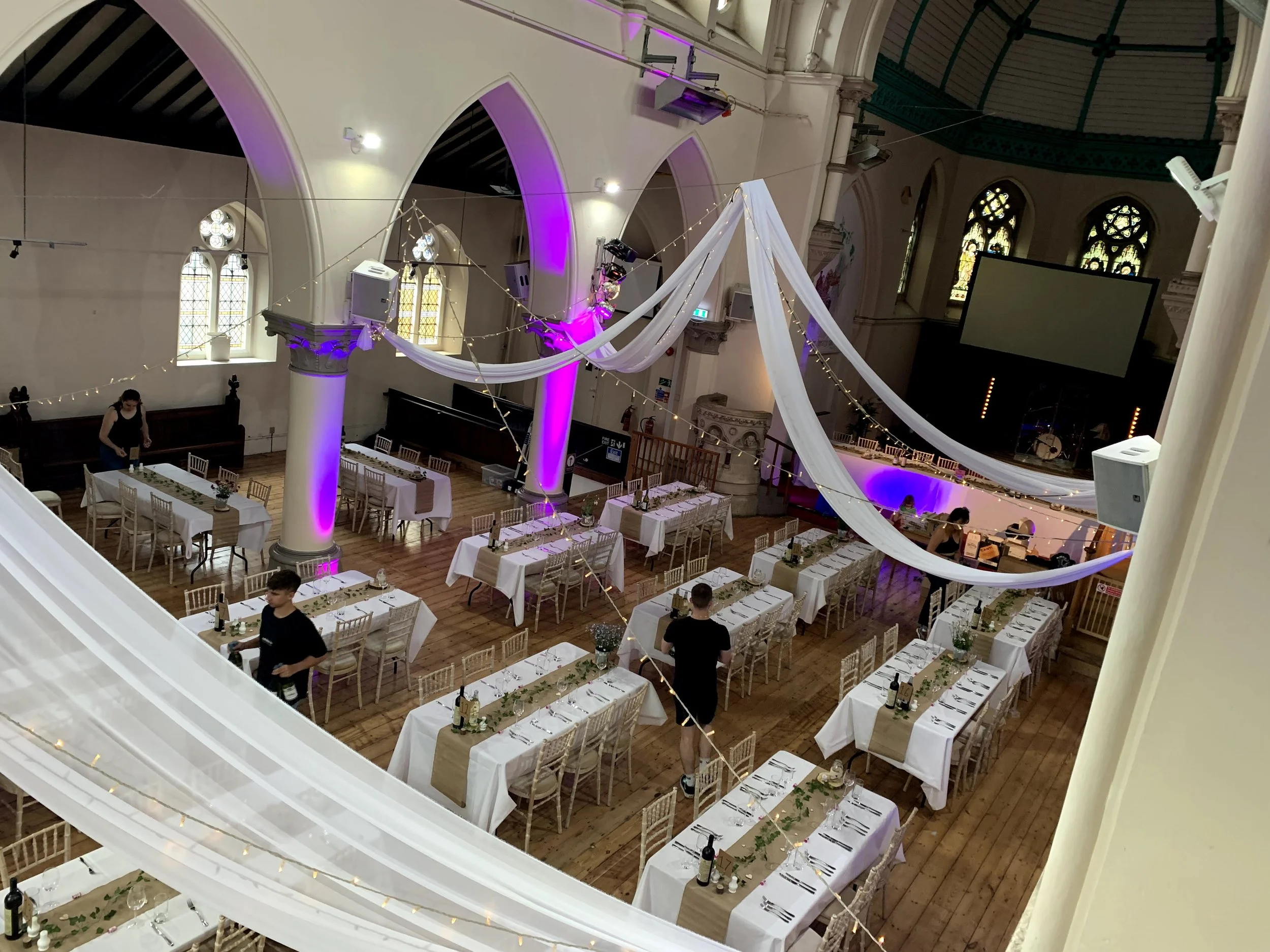Interior of a decorated banquet hall with white tables, chairs, and drapery, illuminated with purple lighting.