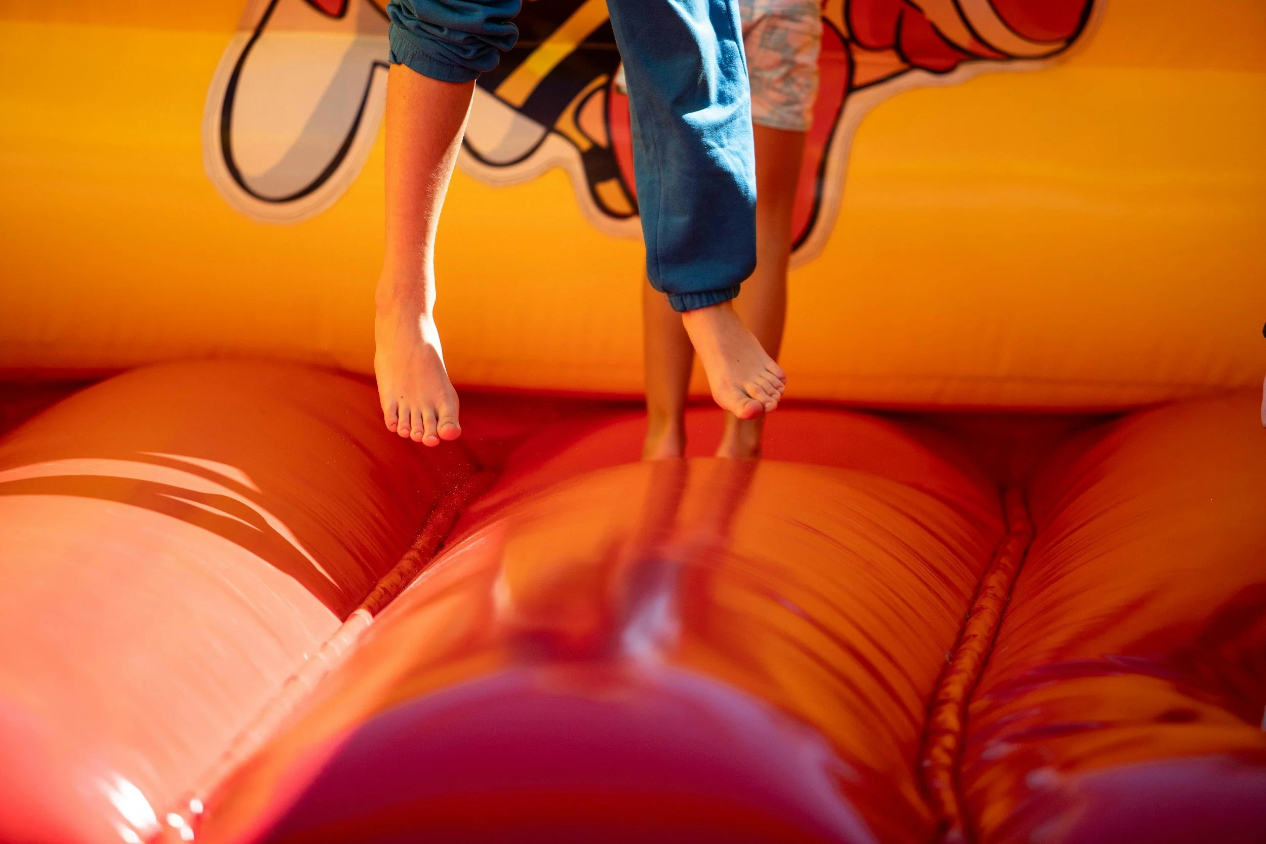 Child wearing blue pants and a gray shirt jumping on a bright orange inflatable bounce house, which has colorful graffiti-style artwork on the side.