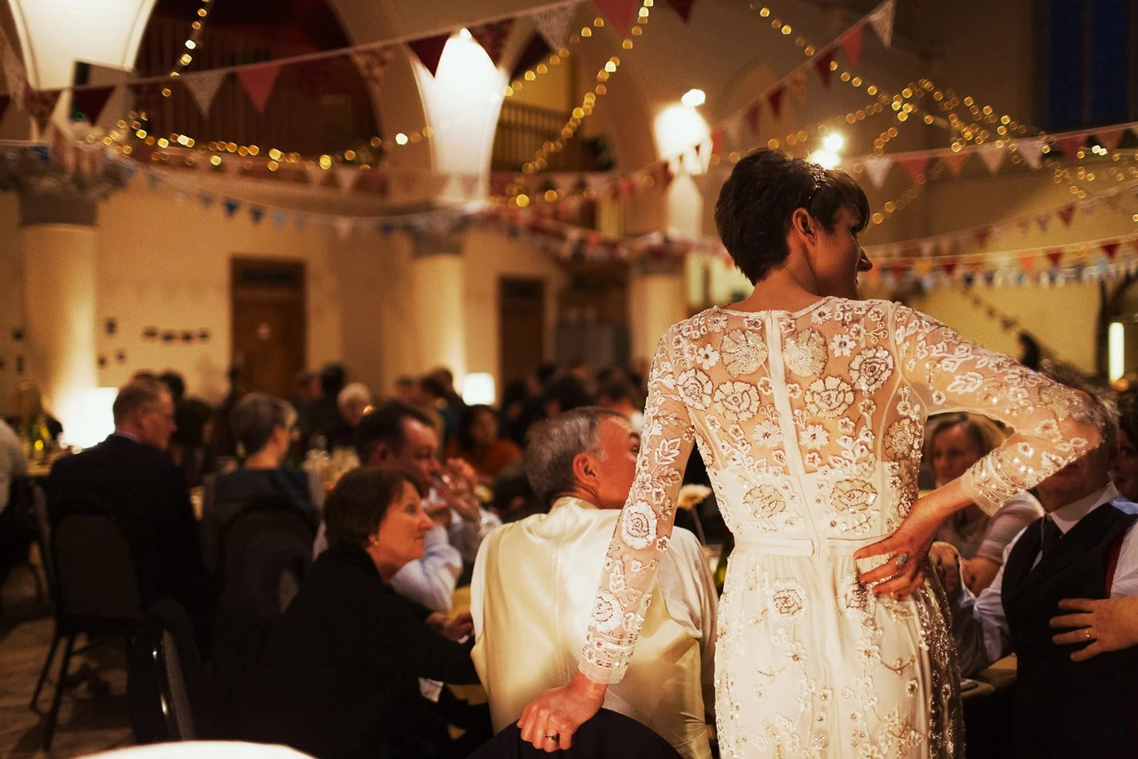 A woman in a white lace dress stands with her hand on her hip, surrounded by seated guests at a decorated indoor event with hanging banners and string lights.