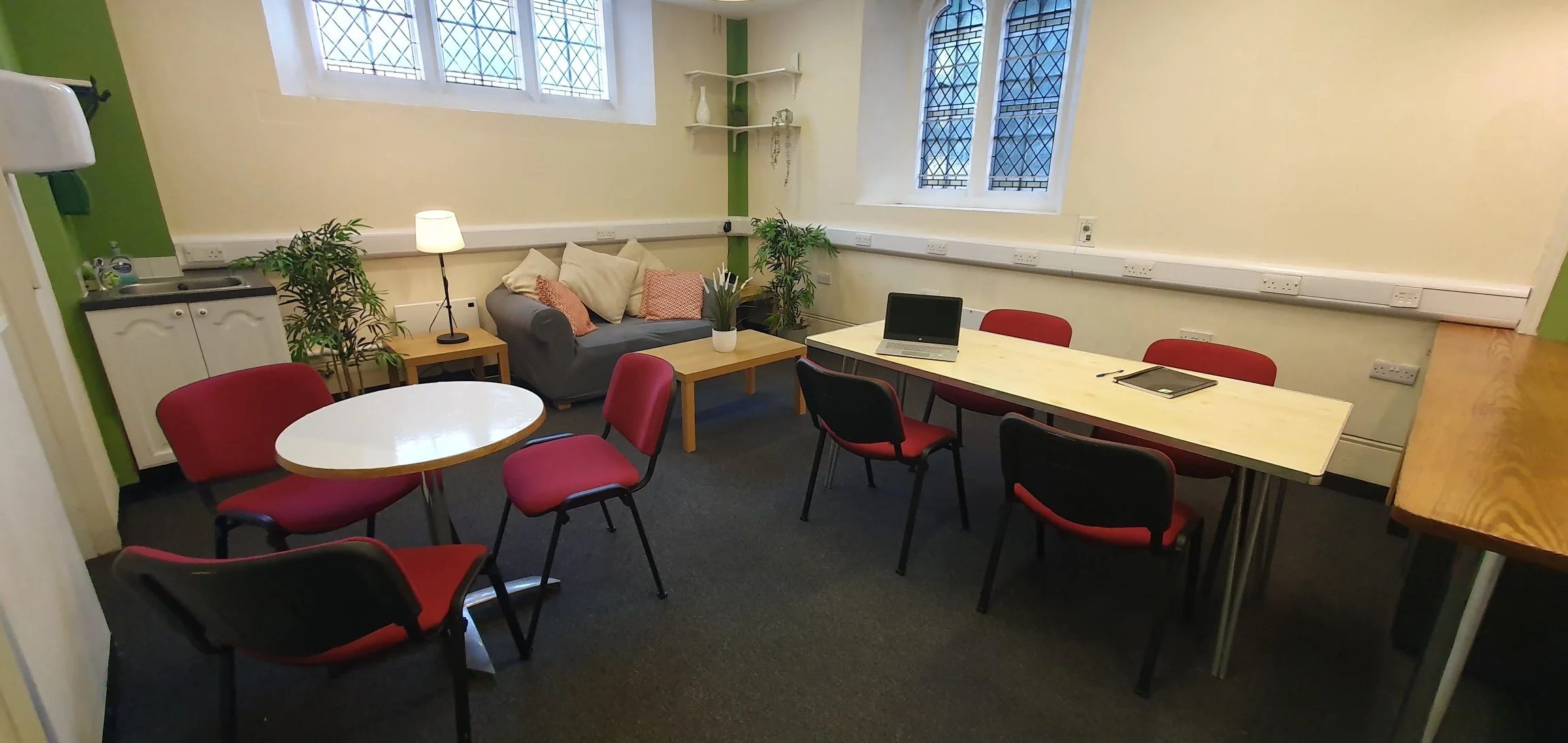 A cozy meeting or lounge room with a round table and red chairs, a long table with laptops and notebooks, a sofa with cushions, potted plants, a small kitchenette, and tall windows with decorative grid patterns.