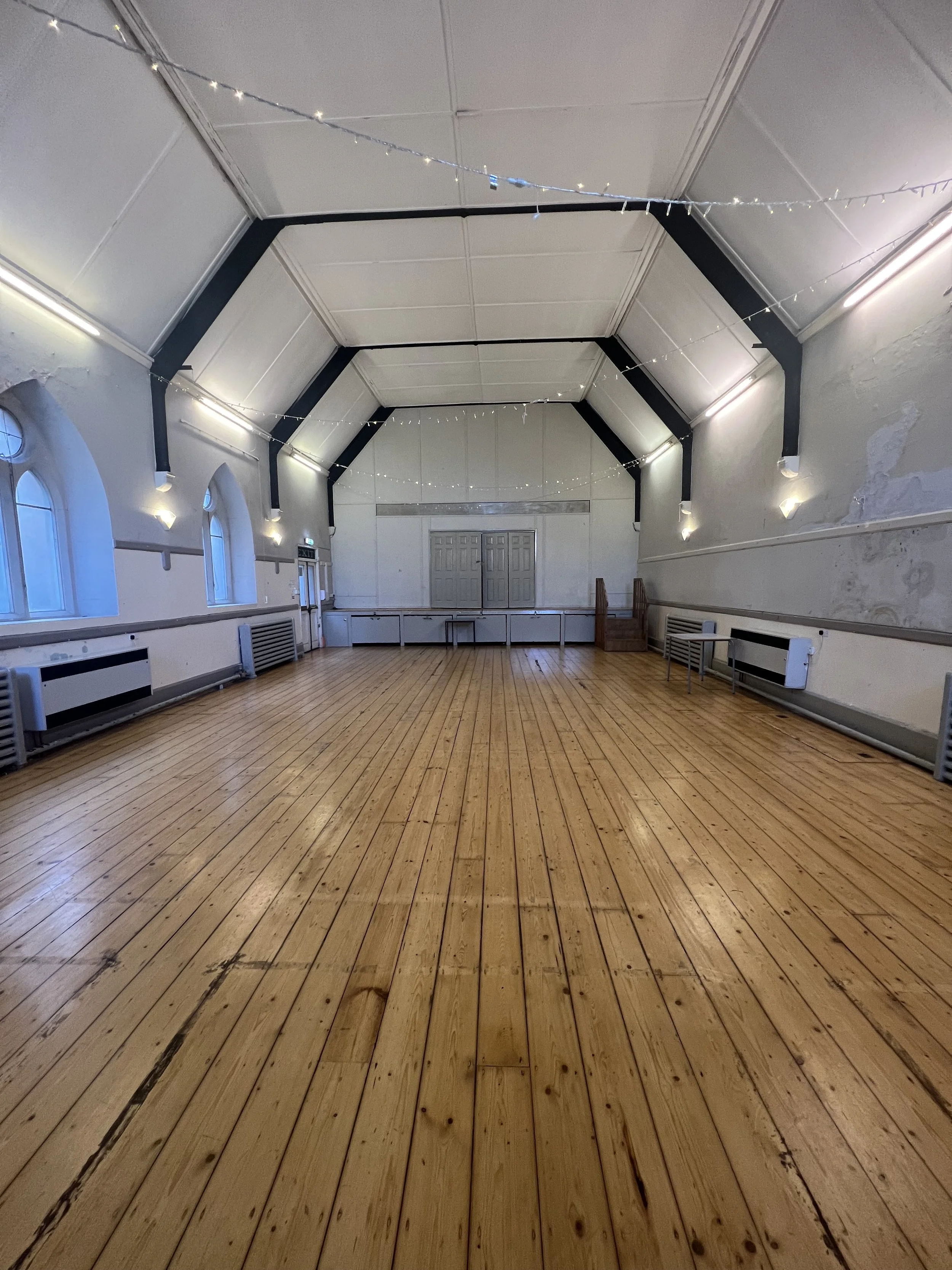 Empty large hall with wooden floors, white walls, arched windows, string lights, and stage at the far end.
