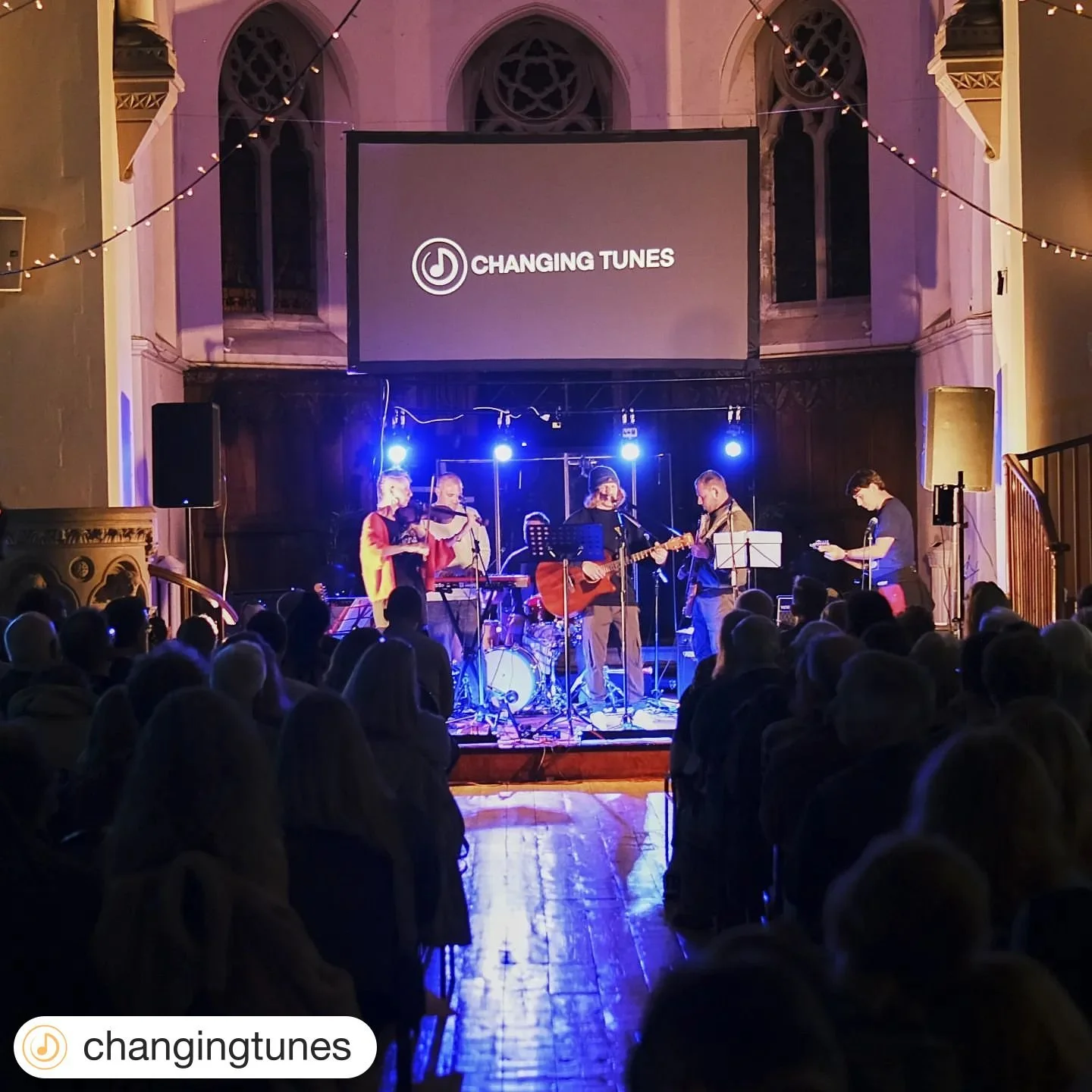 Band performing live on stage at a concert with an audience in front, inside a venue with high arched windows and string lights, with a large screen above displaying the logo and text 'Changing Tunes.'