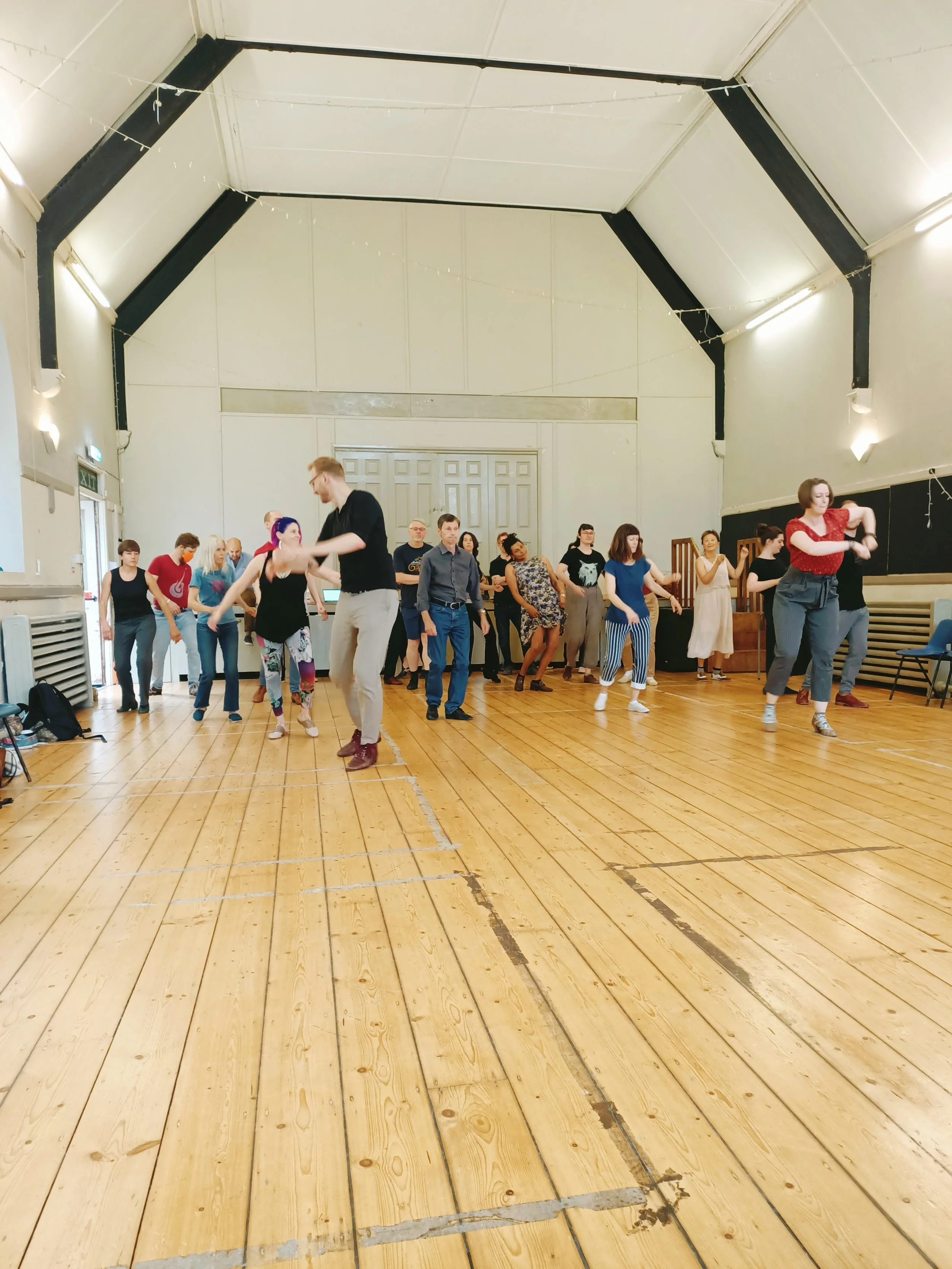 Group of people dancing and socializing in a large, wooden-floored hall, with some individuals actively dancing while others observe.