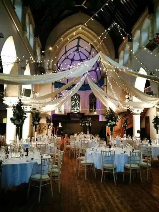 Elegant event hall decorated with white drapery, string lights, floral centerpieces, and round tables with white tablecloths, suited for a wedding or formal celebration.