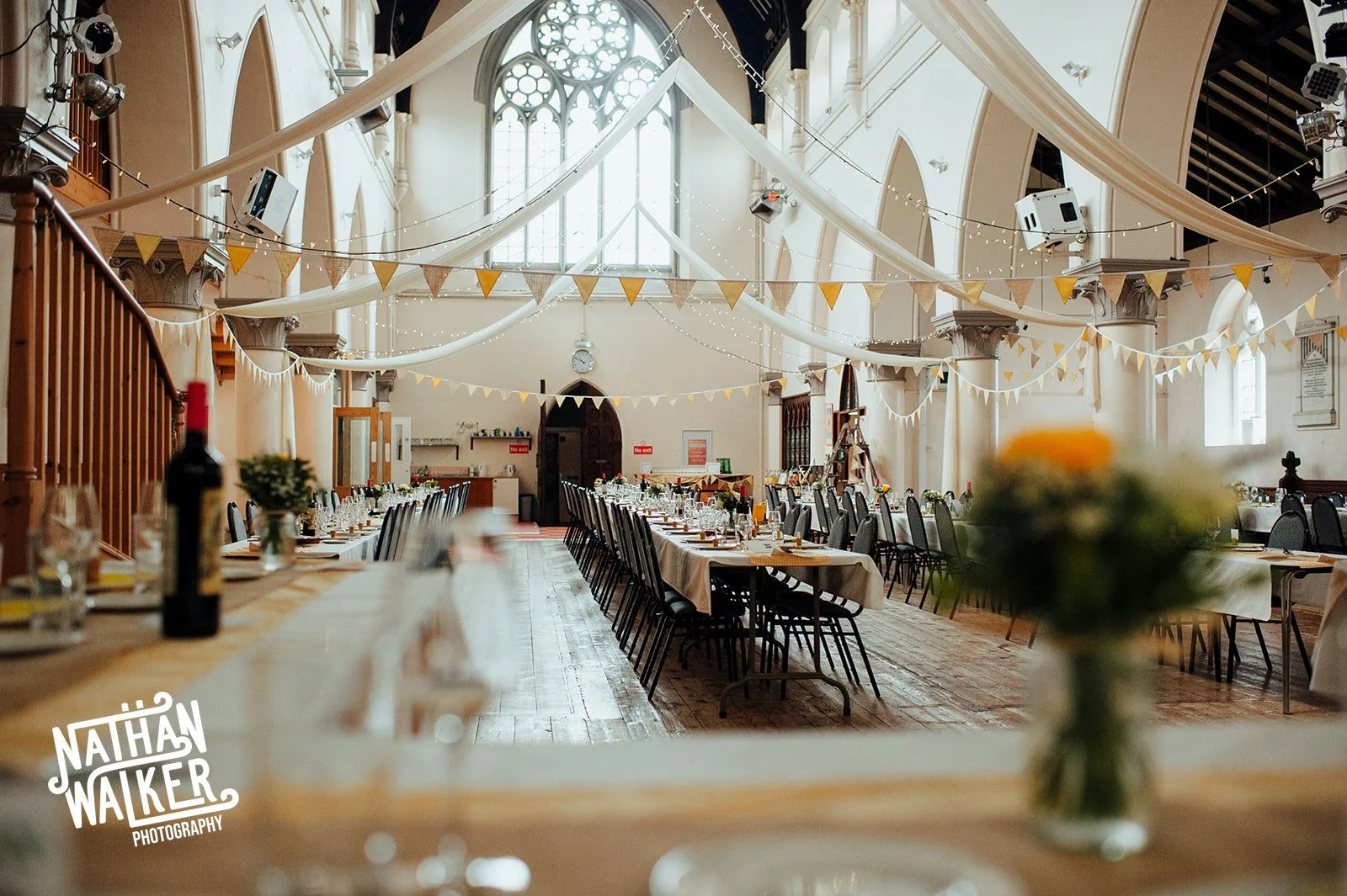 Long banquet tables set with white tablecloths and glassware inside a large, decorated hall with high arched windows, hanging string lights, and bunting banners.