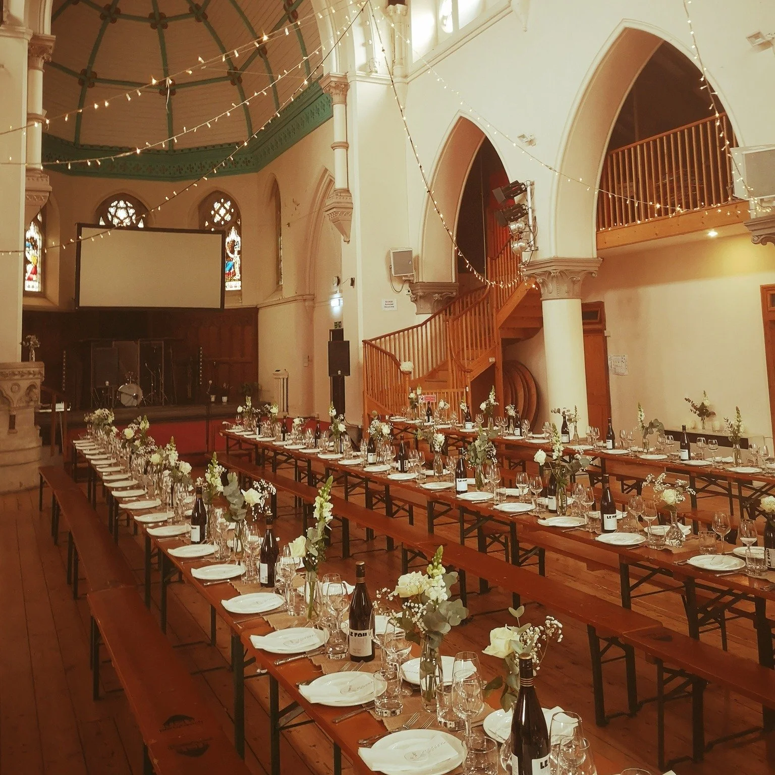 Long banquet tables set with white plates, wine glasses, cutlery, and floral centerpieces inside a decorated church or hall with wooden staircases and string lights.