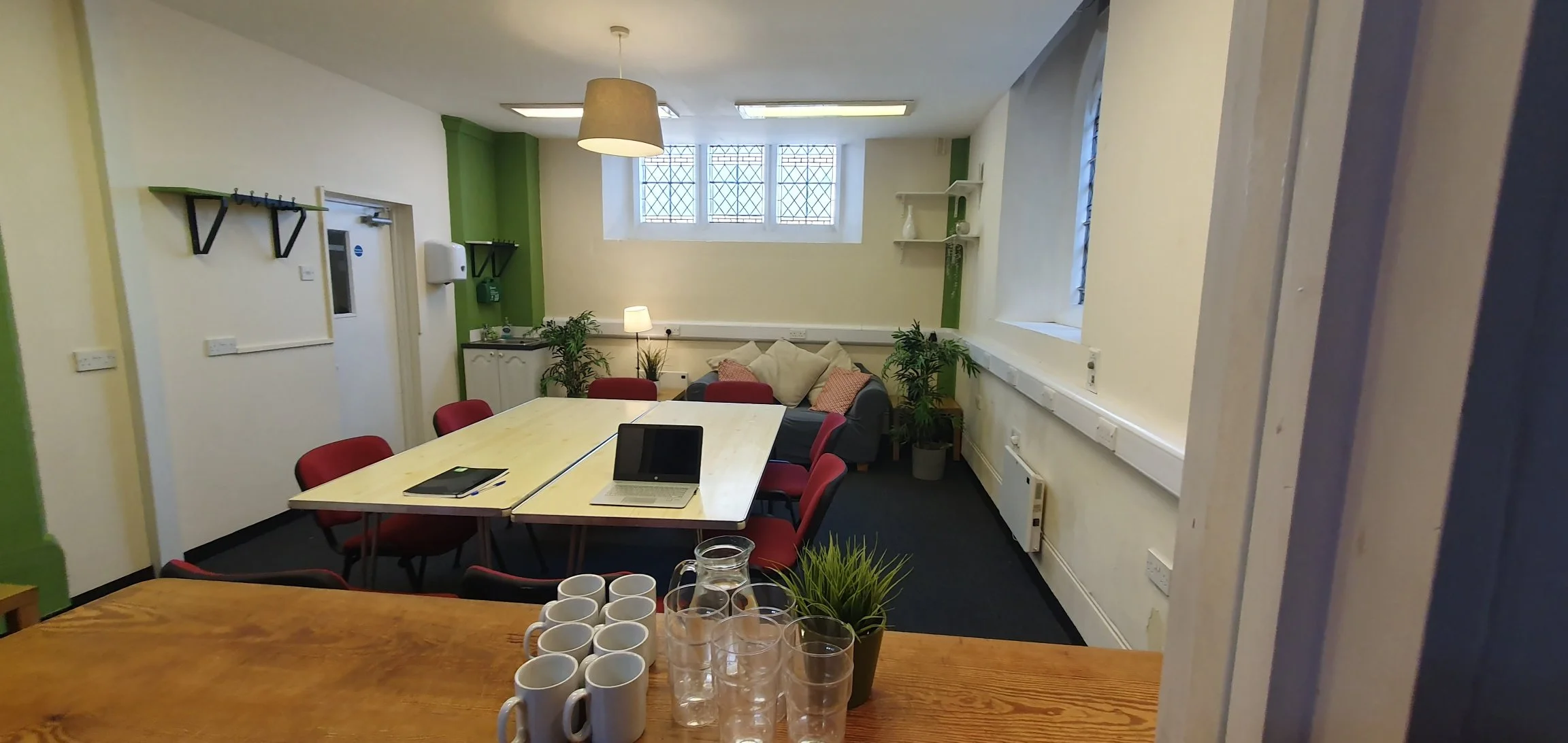 A conference room with a large table, red chairs, a sofa with pillows, plants, a small kitchenette, and windows with bars.