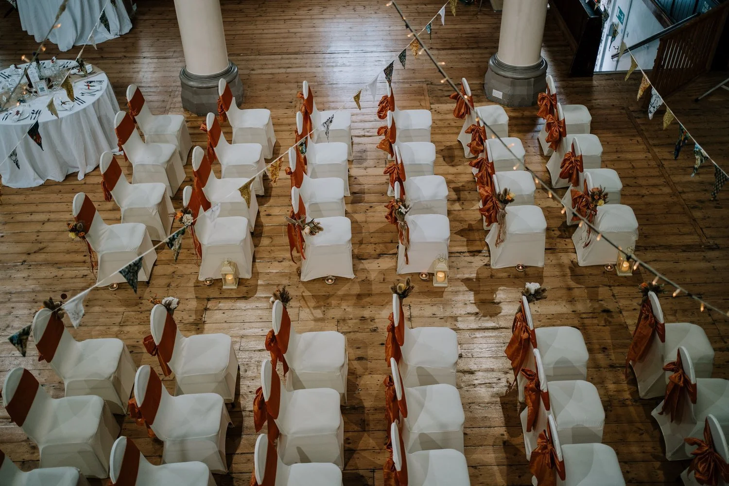 Empty chairs decorated with rust-colored sashes and dried floral arrangements, arranged in rows for a wedding or event in a wooden hall with columns and bunting.