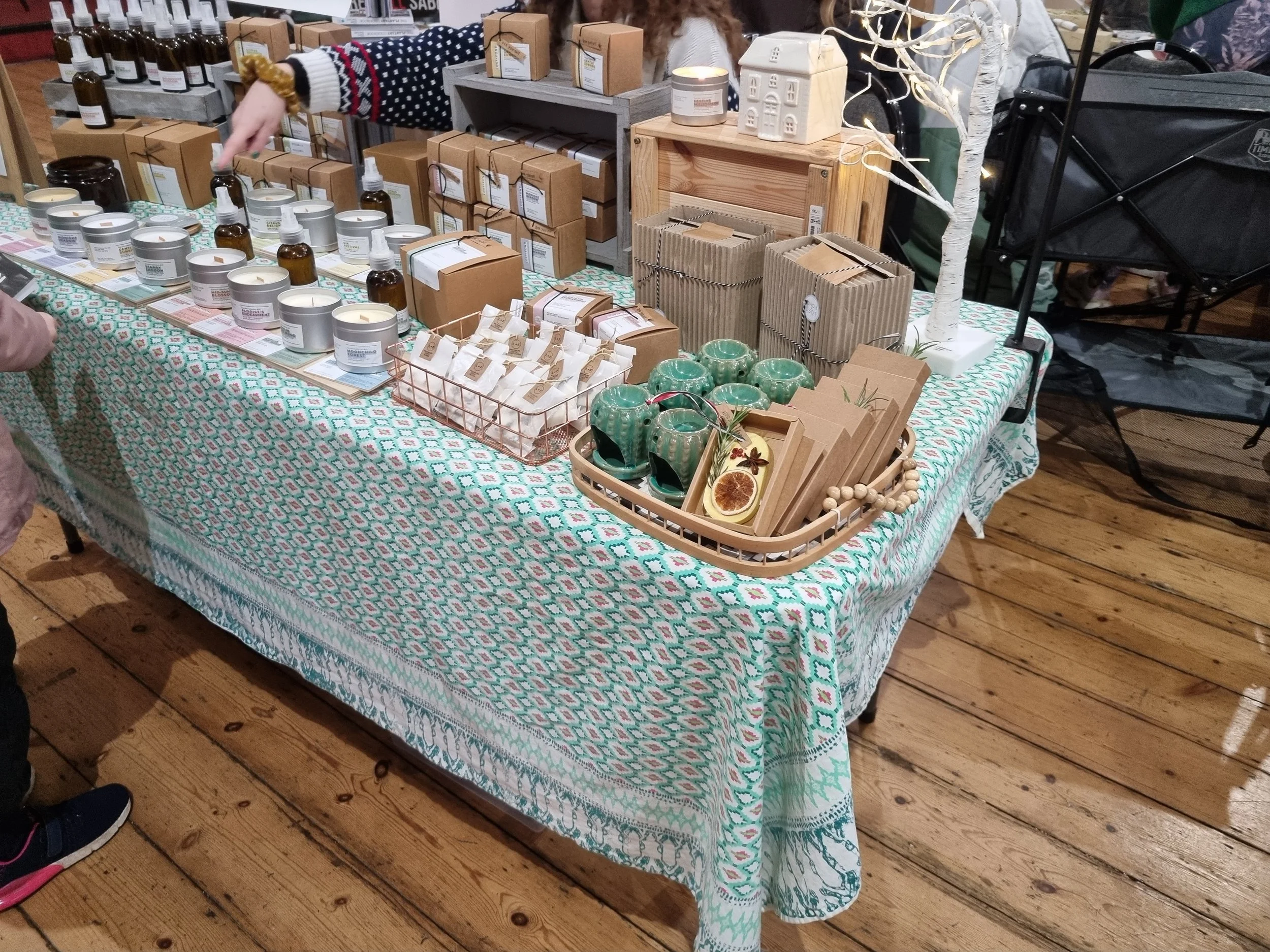 A table covered with a colorful patterned cloth displays various candles, herbal ingredients, and home decor items at a market stall.