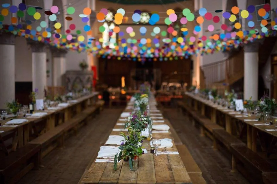 Long wooden banquet table decorated with greenery and white napkins, under a ceiling with colorful hanging paper lanterns, in a rustic-style event hall