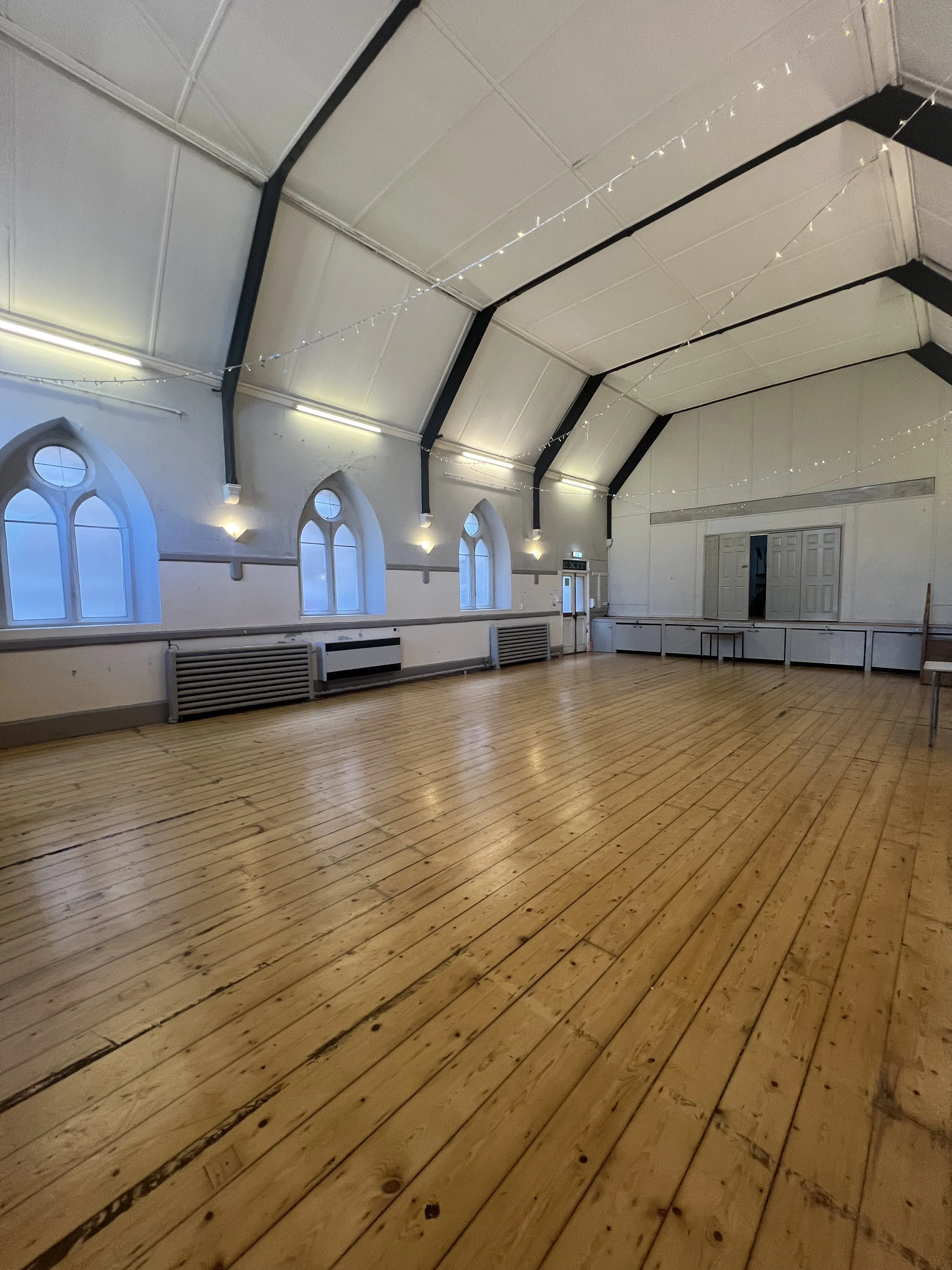 Empty event hall with wooden floor, arched windows, white walls, string lights, and ceiling beams.