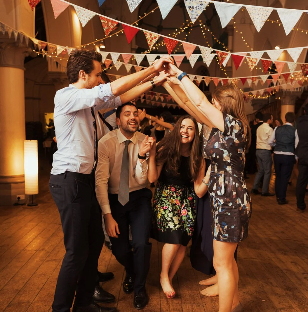 A group of five young adults dancing and enjoying themselves at a festive party with decorative bunting and string lights overhead.