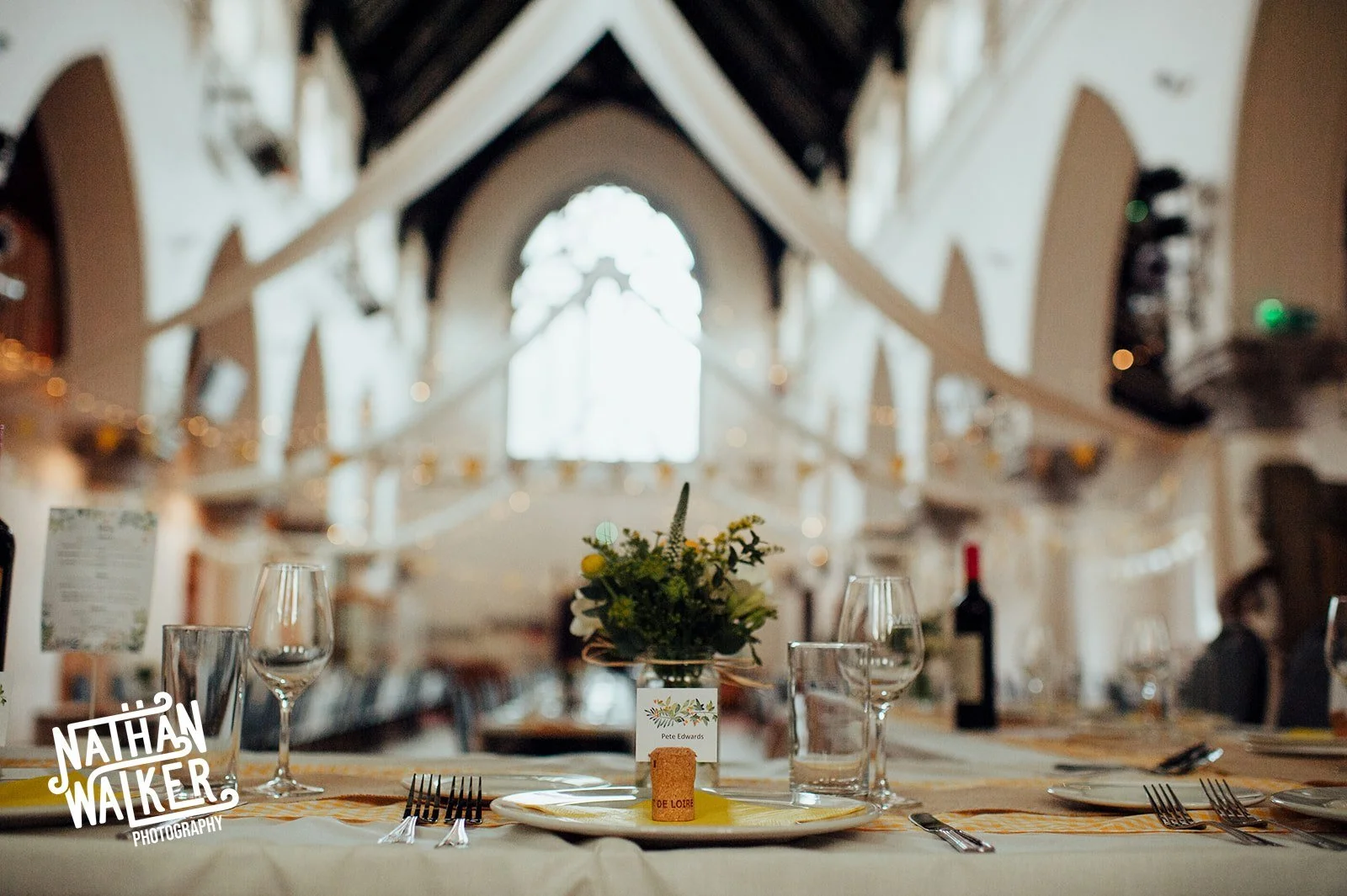 Table setting with a centerpiece flower arrangement, glasses, plates, and cutlery in a decorated hall with high arched ceiling and large window in the background.