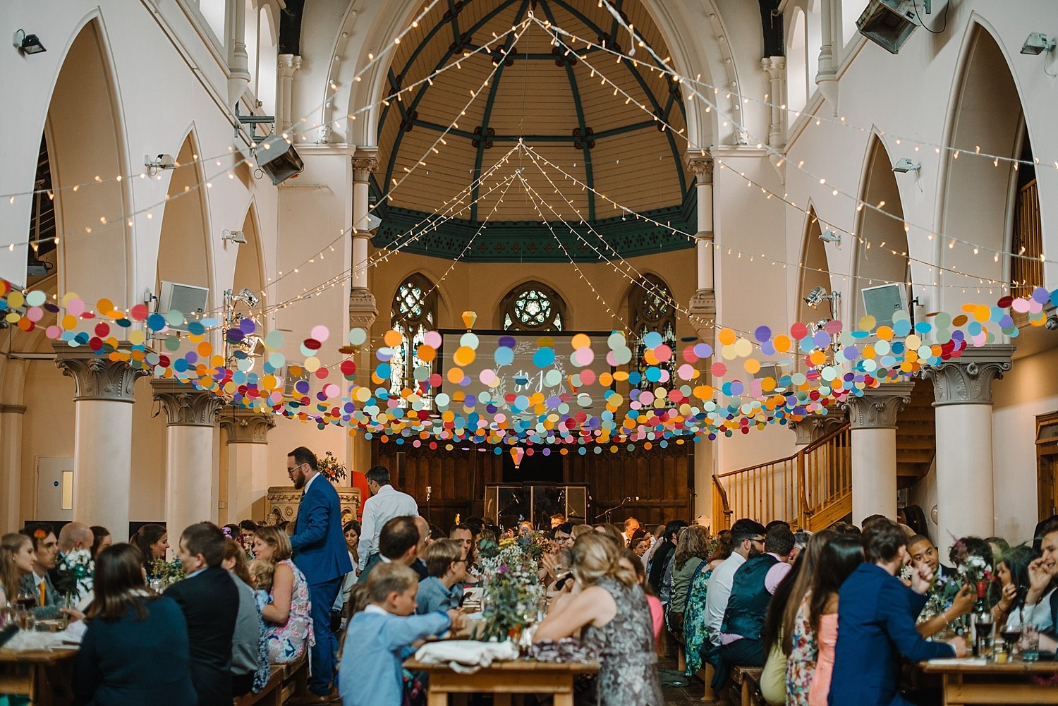Indoor celebration in a decorated venue with hanging string lights and colorful paper circles, guests seated at long tables, and a stage at the back.