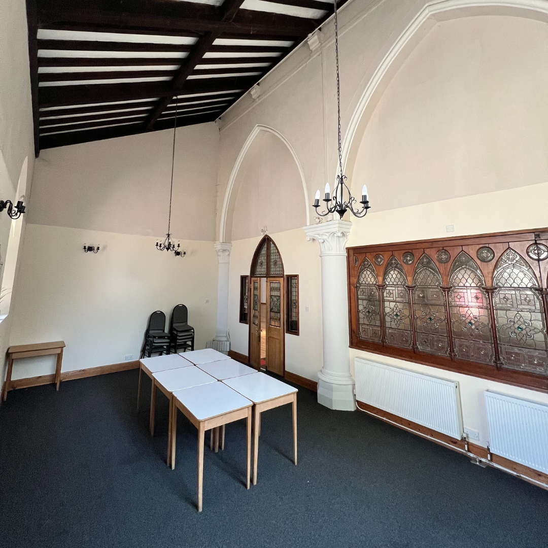 Empty room with high vaulted ceiling, black chandeliers, white walls, wooden accents, and a row of stacked black chairs near the corner.