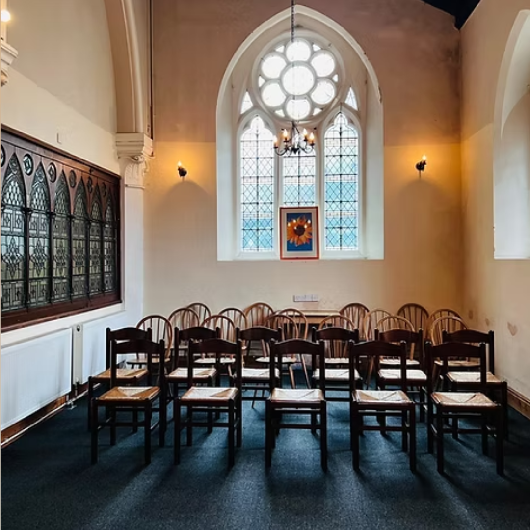 Empty room with wooden chairs arranged in a U-shape, large stained glass window, and a chandelier, resembling a church or historic building interior.