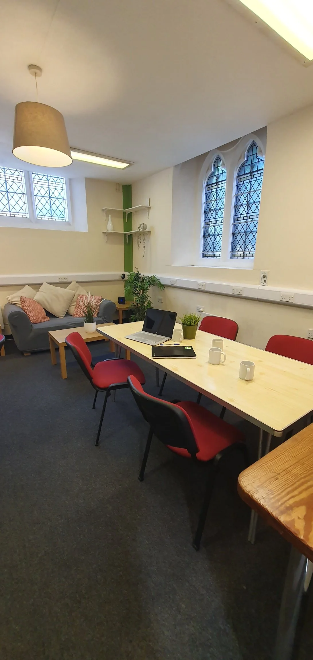 A cozy meeting room with a wooden table, red and black chairs, a gray sofa with pillows, potted plants, a laptop, coffee mugs, and large windows with decorative stained glass.