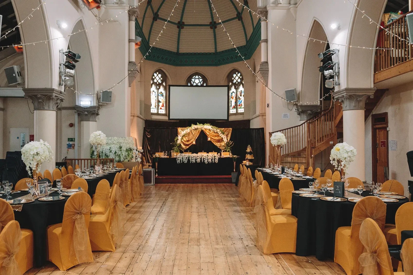 Elegant banquet hall decorated for a wedding reception with round tables covered in black tablecloths and gold chairs with bows, floral centerpieces, a decorated head table, and a stage with drapery, flowers, and a cake, inside a historic building wi
