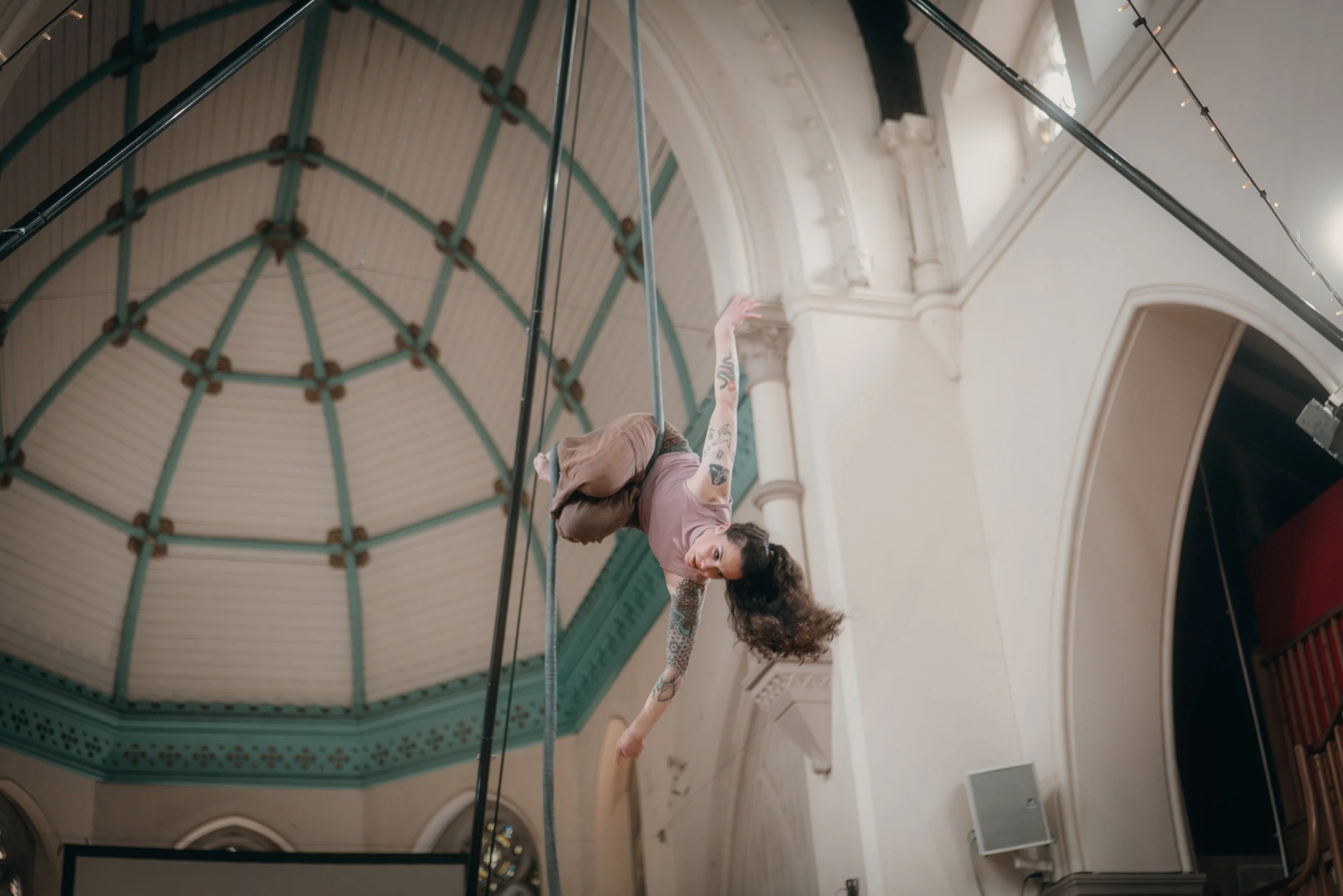 A woman with tattoos and curly hair, wearing a pink shirt and brown pants, is performing a dance or acrobatic move on a hanging pole inside a spacious, ornate hall with high ceilings and intricate architectural details.