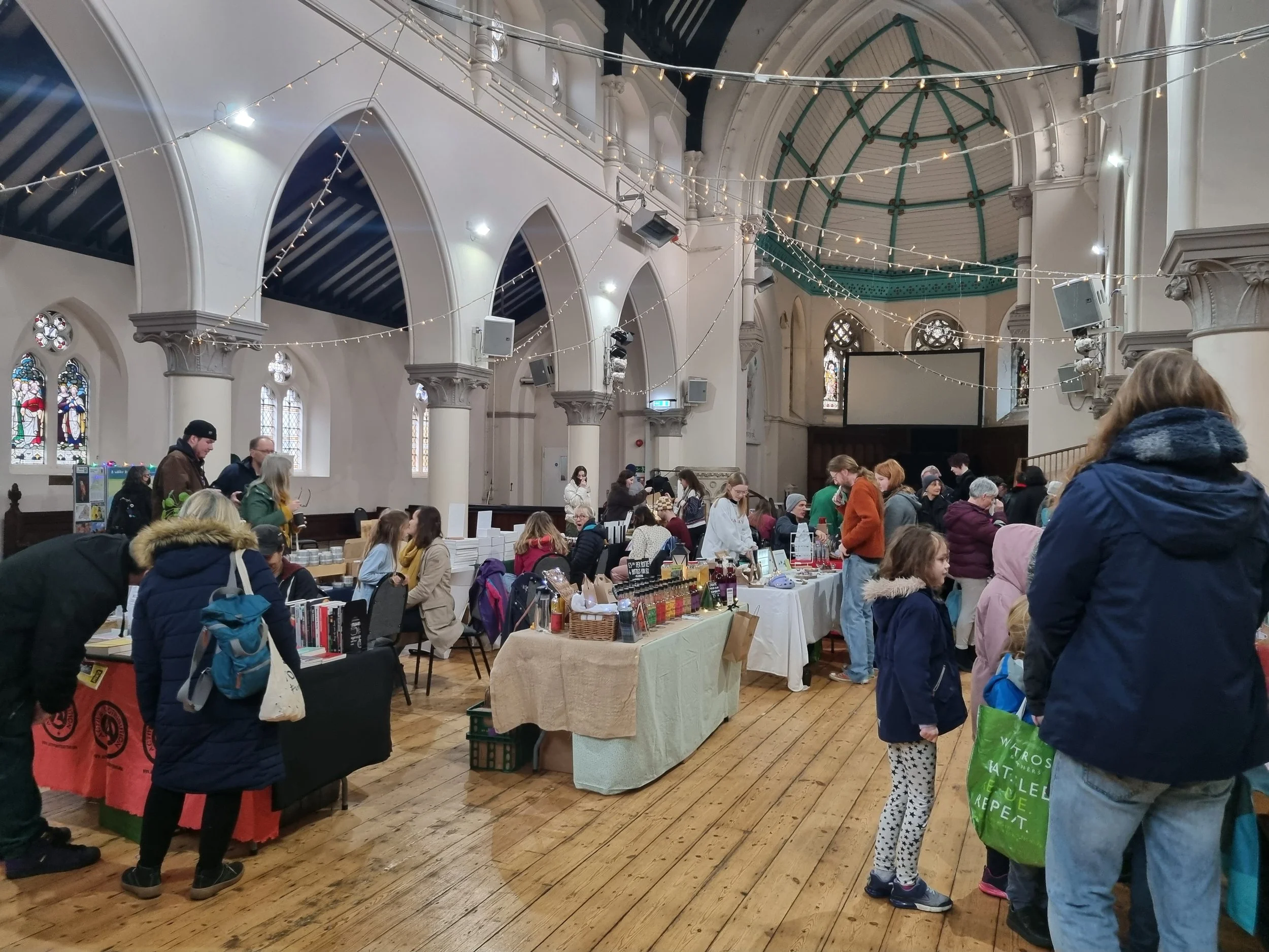 A busy indoor market inside a church with high arched ceilings, stone columns, and stained glass windows. People browse tables with books, crafts, and baked goods, with string lights hanging overhead.