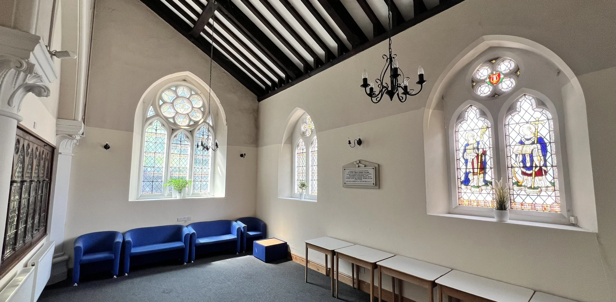 Interior of a church or chapel with stained glass windows, a black chandelier, some blue chairs, and light-colored walls.