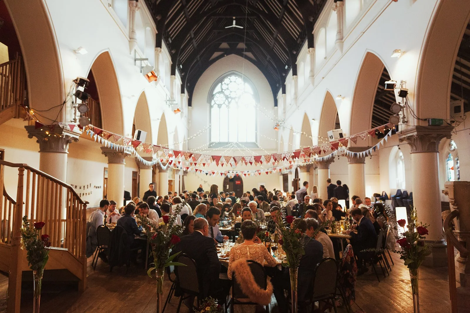 A large gathering of people seated at decorated tables inside a church or hall during a celebration or event, with bunting hanging from the ceiling and large windows at the back.