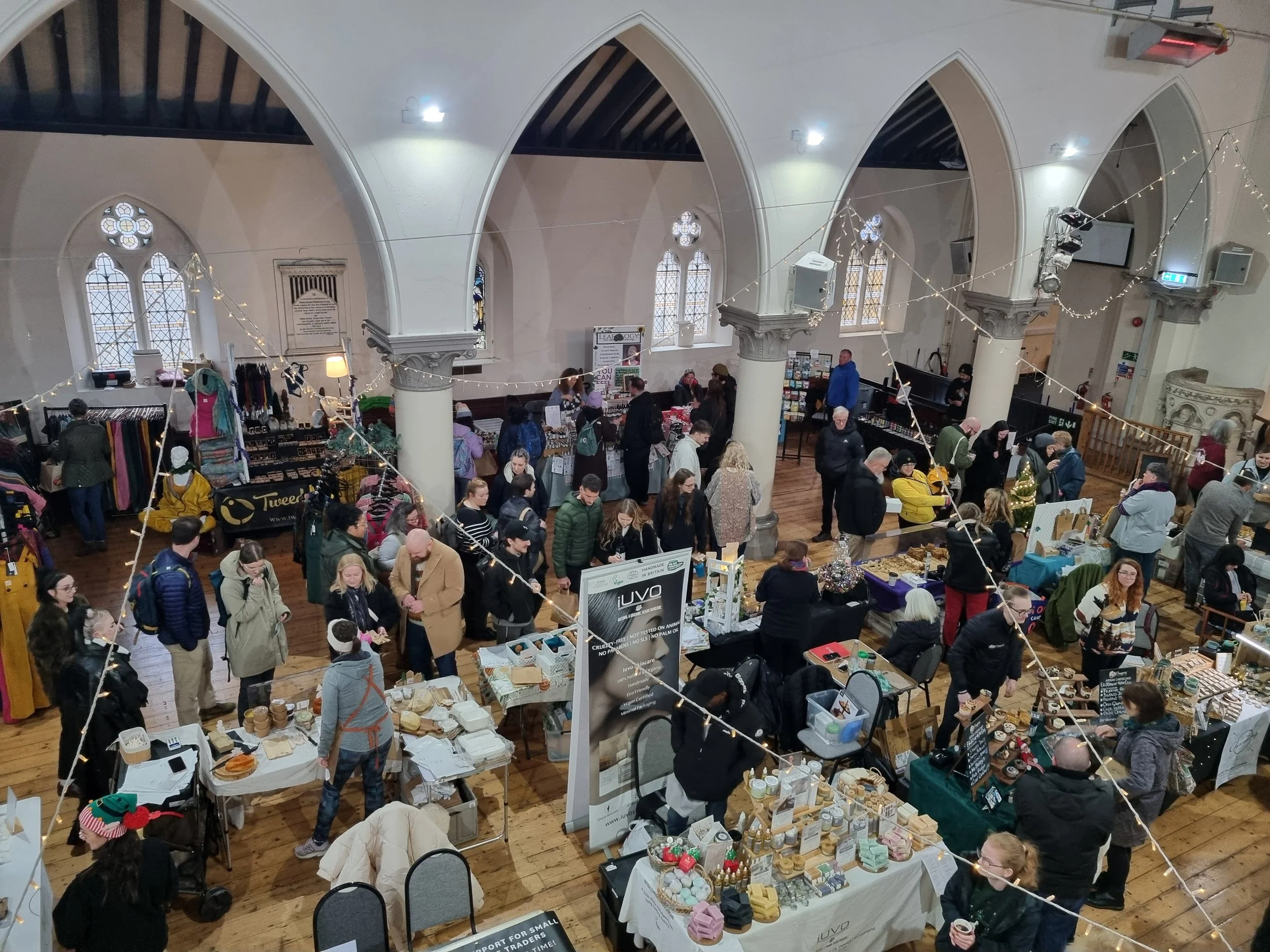Indoor marketplace with various stalls selling handmade items, jewelry, and crafts, inside a church-like building with high arched windows and wooden floors, crowded with shoppers.