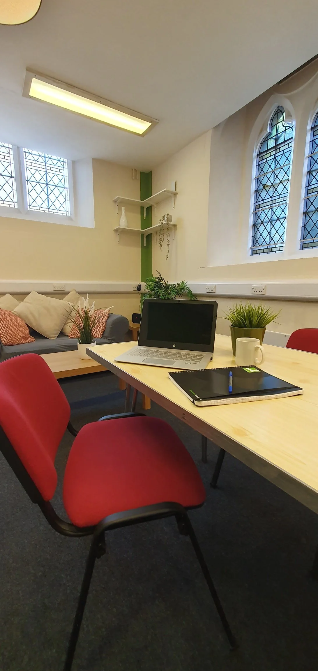 Office meeting room with a wooden table, red chairs, a laptop, notebooks, and plants. In the background, there's a couch with pillows and windows with stained glass.