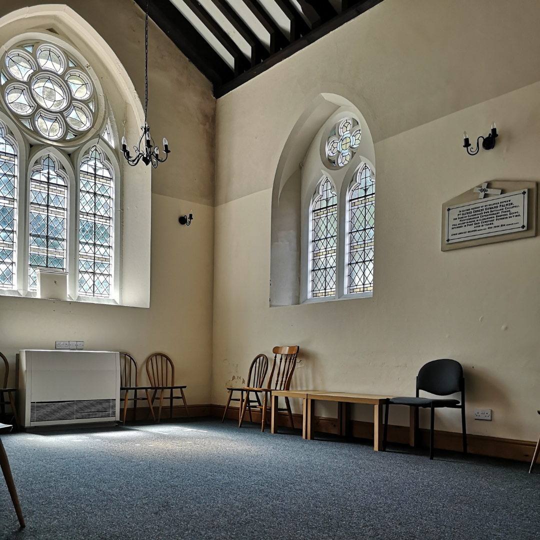 Empty room with tall stained glass windows, wooden chairs, and a black chair, inside a church or chapel.
