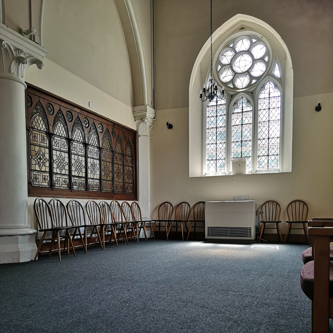 An empty room in a church with a large stained glass window, wooden chairs lined along the wall, a chandelier hanging from the ceiling, and a carpeted floor.