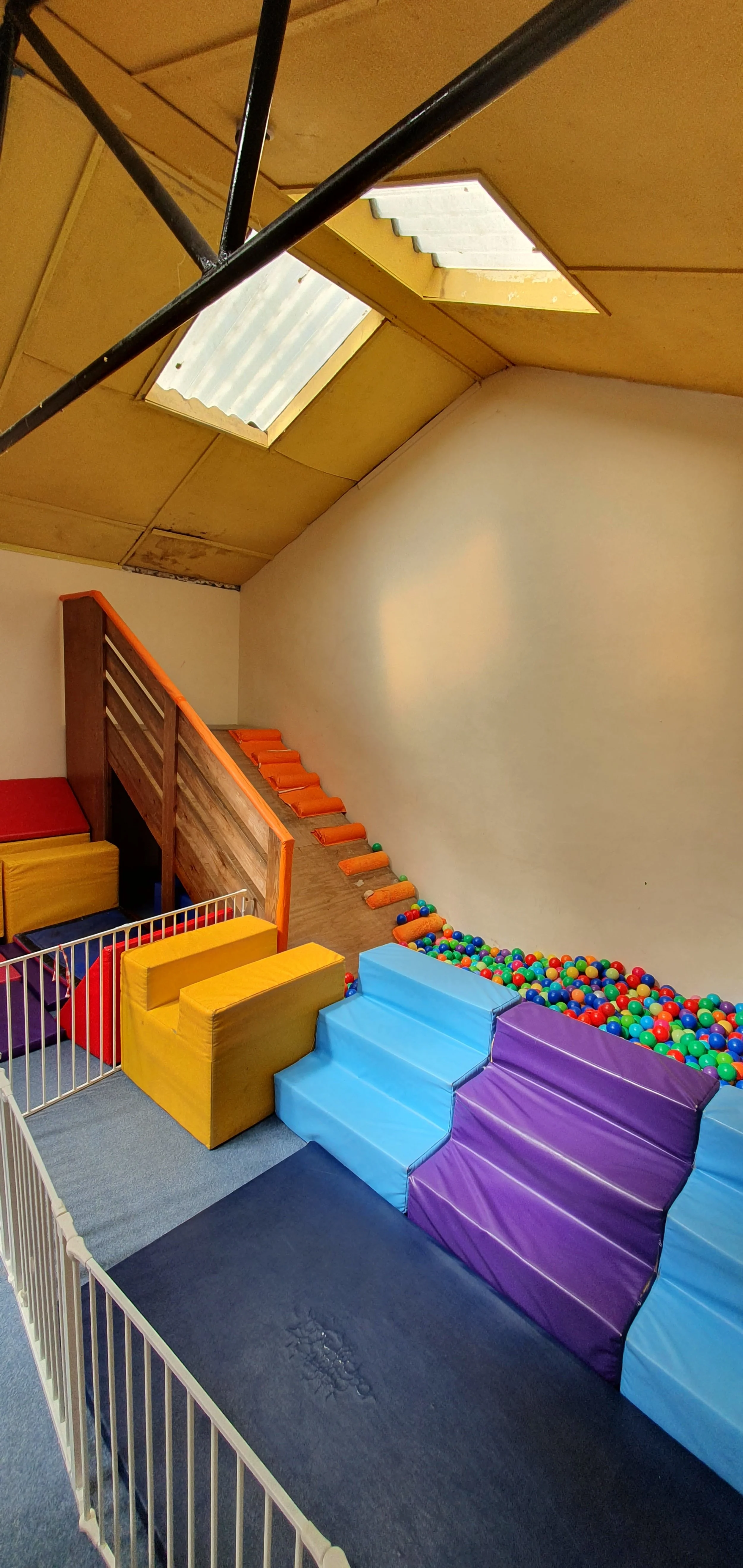 Indoor children's play area with colorful foam blocks, a ball pit filled with multicolored plastic balls, a blue staircase, and a wooden climbing wall with orange footholds, under a ceiling with skylights.